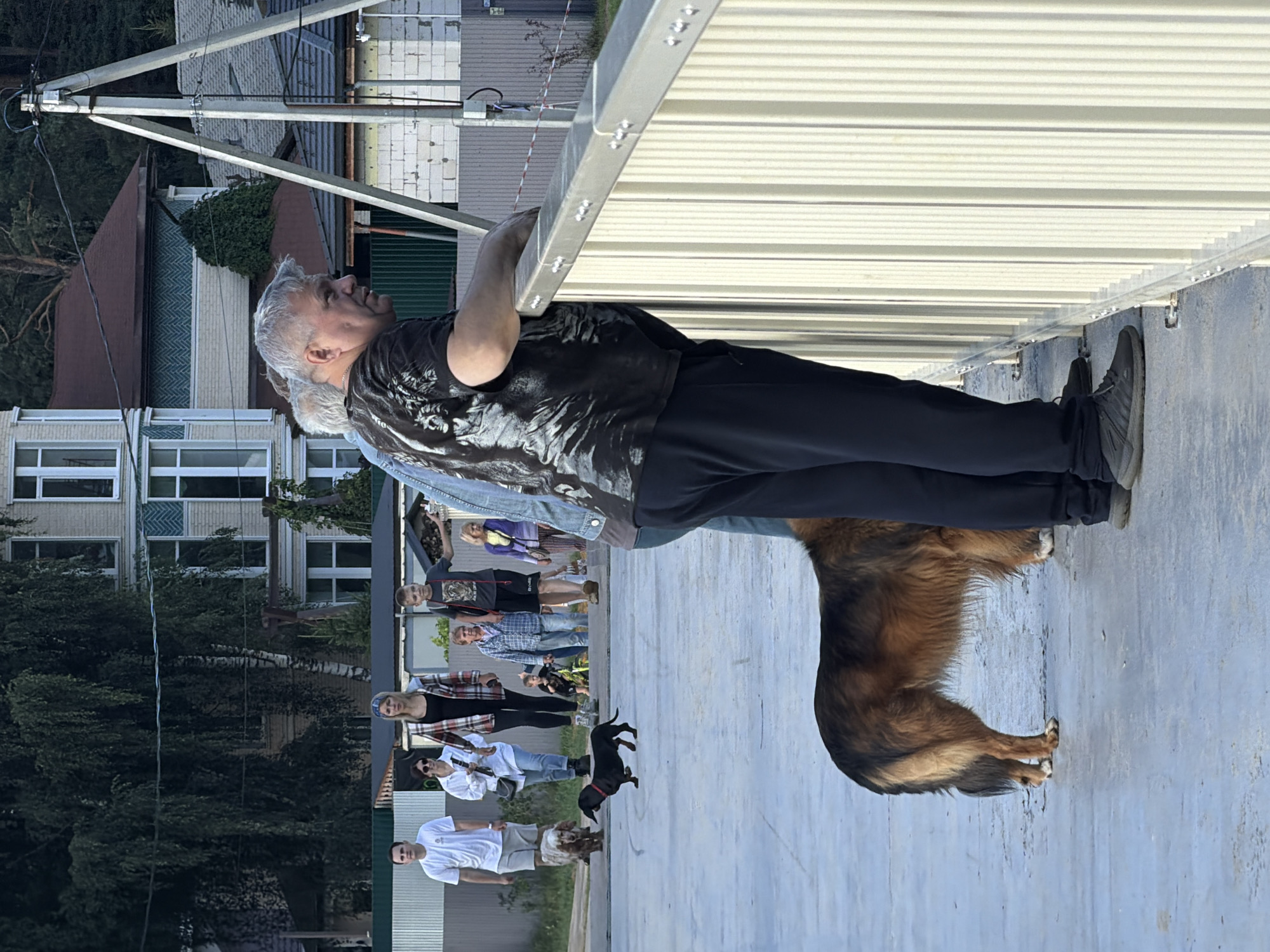In Malakhovka, Russia, a group of people and two dogs are gathered on a paved area. In the foreground, a man with gray hair and a black t-shirt with a white graphic print is leaning on a light-colored metal railing. Next to him, a large, long-haired brown and black dog stands facing left. In the mid-ground, several other people are walking or standing. A man in a white t-shirt and shorts walks towards the left, accompanied by a small, light-colored dog. A woman in a plaid shirt over a black top and jeans walks beside him, holding a bag. Behind them, a boy in a dark t-shirt and shorts, and a younger child on a bicycle, are visible. Further back, a woman in a blue plaid shirt and a woman in a light purple jacket are also present. In the background, several houses with different architectural styles and roof colors are visible, along with trees and a utility pole. The overall lighting suggests it is late afternoon or early evening.