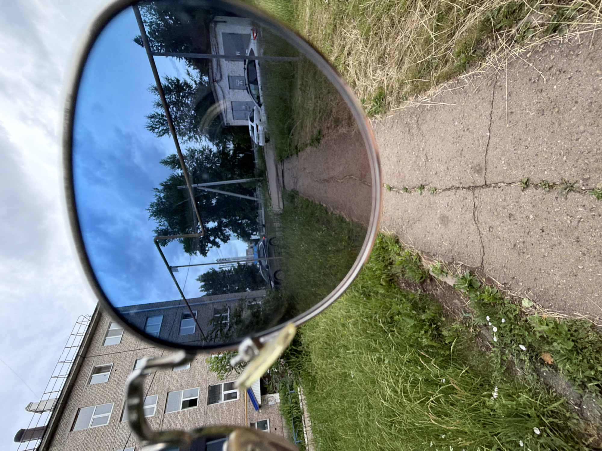 The image depicts a reflective view through a pair of blue-tinted sunglasses. The foreground is dominated by the rim of the sunglasses and their blue lenses, which reflect a scene of an urban courtyard. Reflected in the lenses, a cracked concrete pathway leads away from the viewer, flanked by overgrown grass and weeds. To the left of the pathway, a multi-story brick apartment building with numerous windows is visible. To the right, a row of small garages with closed doors and a white car are reflected. The sky in the reflection is cloudy and overcast. In the real foreground, outside the reflection, the pathway continues, with dry grass and some small white wildflowers growing along its edge. The scene suggests a residential area in a Russian city, likely during the daytime given the lighting. The sunglasses are held in front of the camera, creating a framed perspective of the reflected environment.