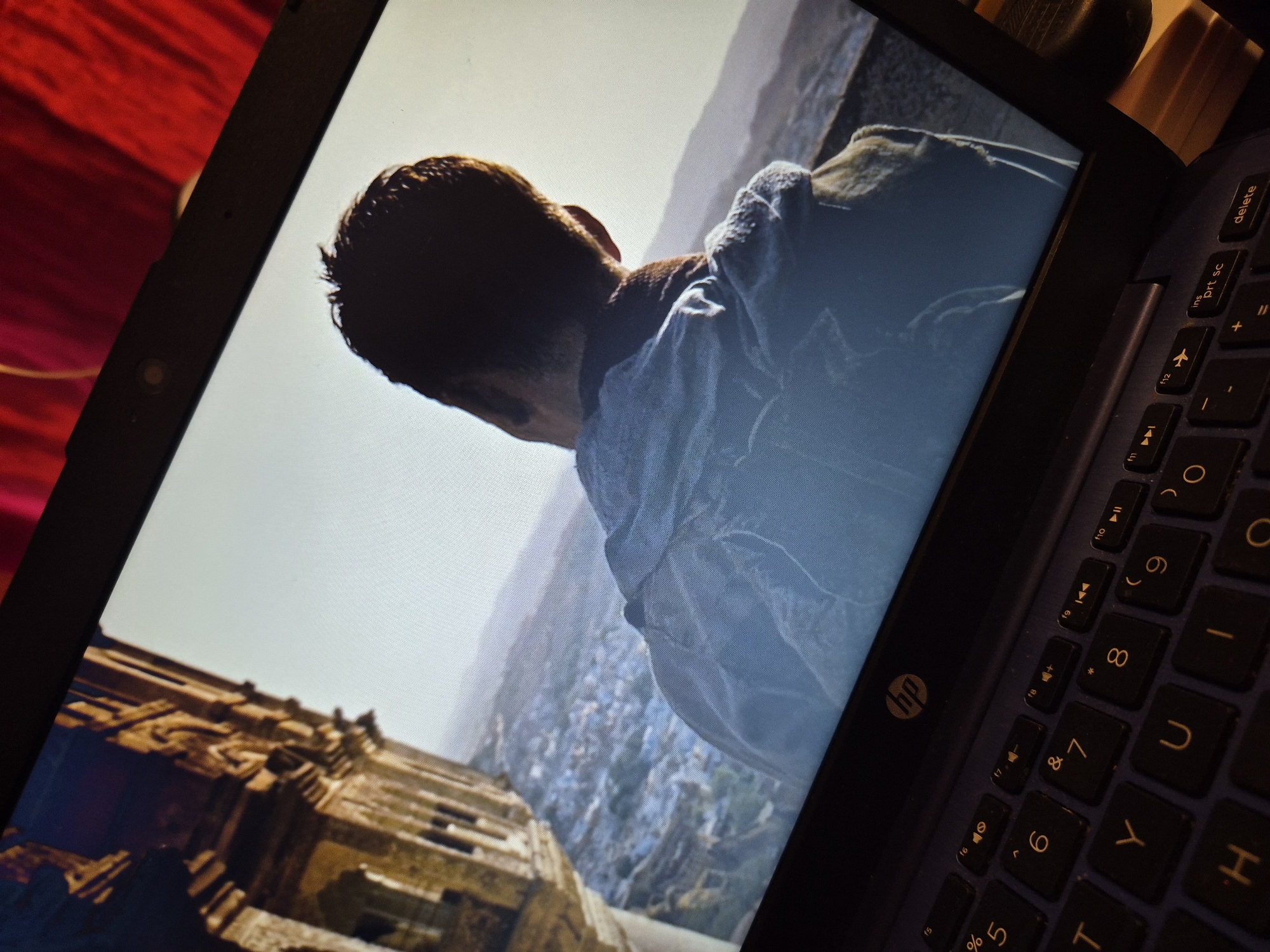 The image shows a laptop screen displaying a scene of a man from behind, looking out over a city. The laptop is dark blue with a black keyboard. The screen displays a person wearing a light-colored jacket, with their back to the viewer. They are looking out at a landscape that appears to be a city with buildings and mountains in the distance. On the left side of the screen, there is a stone structure that resembles an ancient building or ruin. The overall lighting suggests it is daytime. The lower part of the image shows a close-up of the laptop's keyboard, with the "hp" logo visible on the top edge of the laptop. The background behind the laptop is out of focus and appears to be a red fabric. There is no indication of the location being in Sandwell, United Kingdom.