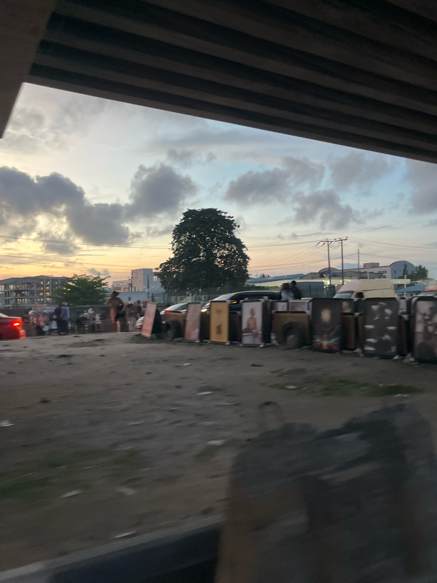 This image depicts a street scene at dusk in Ajah, Nigeria, likely near a market or roadside stall. A thick, dark structure, possibly an overpass or bridge, looms over the upper portion of the frame. The sky is filled with dramatic, textured clouds, suggesting the end of the day or an approaching weather system.
The ground appears to be unpaved, with debris scattered across it. In the mid-ground, a collection of large, framed artworks or posters are displayed upright, lining what appears to be a roadside. Some of these artworks feature portraits. Several vehicles, including cars and what could be a van, are present, some parked and others possibly in motion, indicated by motion blur.
Several individuals are visible in the scene, engaged in various activities. Some are standing around the artworks, possibly as vendors or potential customers. Others are walking or gathered in small groups. The overall impression is of a bustling, informal commercial area active during the twilight hours. The presence of buildings in the background suggests an urban or semi-urban environment.