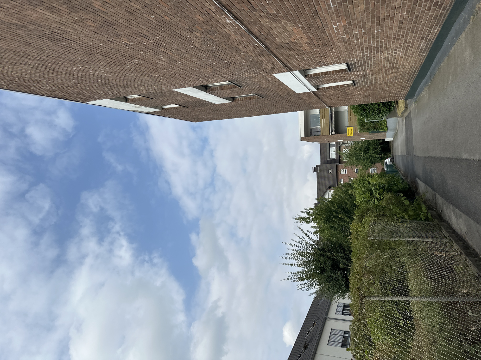 The image captures a narrow, paved alleyway between buildings in Krefeld, Germany. On the right, a tall, reddish-brown brick building dominates the foreground, angled towards the viewer, with several rectangular white-framed windows visible. A thin, silver conduit runs horizontally along the brick wall. To the left of the alley, a chain-link fence runs along a green hedge, beyond which are residential buildings with dark tiled roofs and a white building with horizontal blinds. Further into the distance, other multi-story buildings are visible, including one with a balcony and a yellow sign that reads "Achtung! Betreten des Geländes ist strengstens verboten." In the background, a white car is parked. The sky above is blue with scattered white clouds. The scene appears to be a typical urban residential environment under daylight.