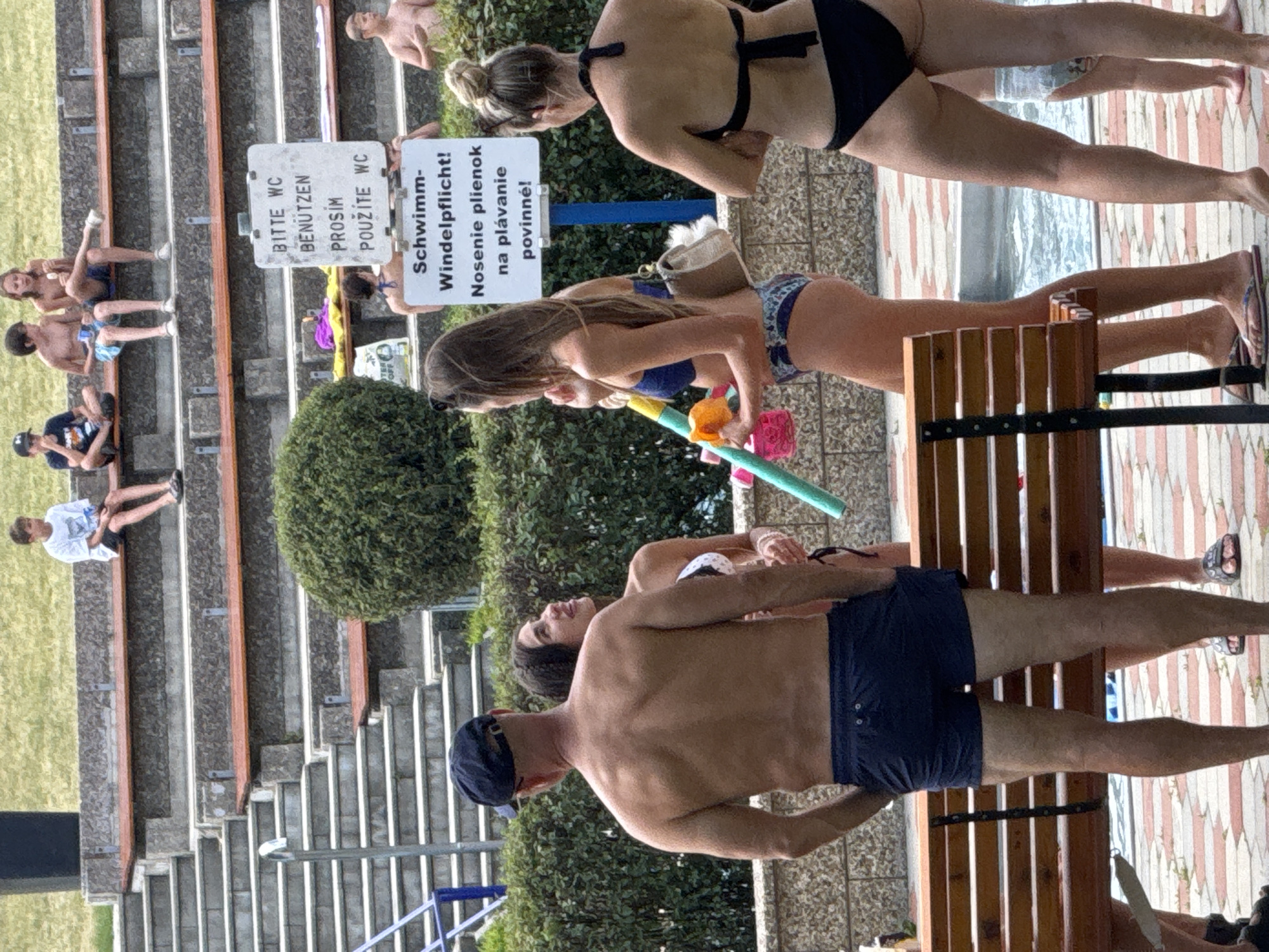 The image shows an outdoor scene at a public swimming pool area in Hainburg an der Donau, Austria. In the foreground, a man wearing dark blue swimming trunks and a dark baseball cap stands with his hands in his pockets, facing away from the camera. To his right, a woman in a blue bikini is talking to another woman wearing a black bikini. Behind them, a white sign with blue text is posted on a pole. The sign has text in German and Slovak, indicating rules for the swimming pool. Further back, several children and adults are seated on tiered stone bleachers. Some children are eating ice cream. The environment includes a swimming pool, paved ground with a red and white pattern, a wooden bench, and lush green bushes.