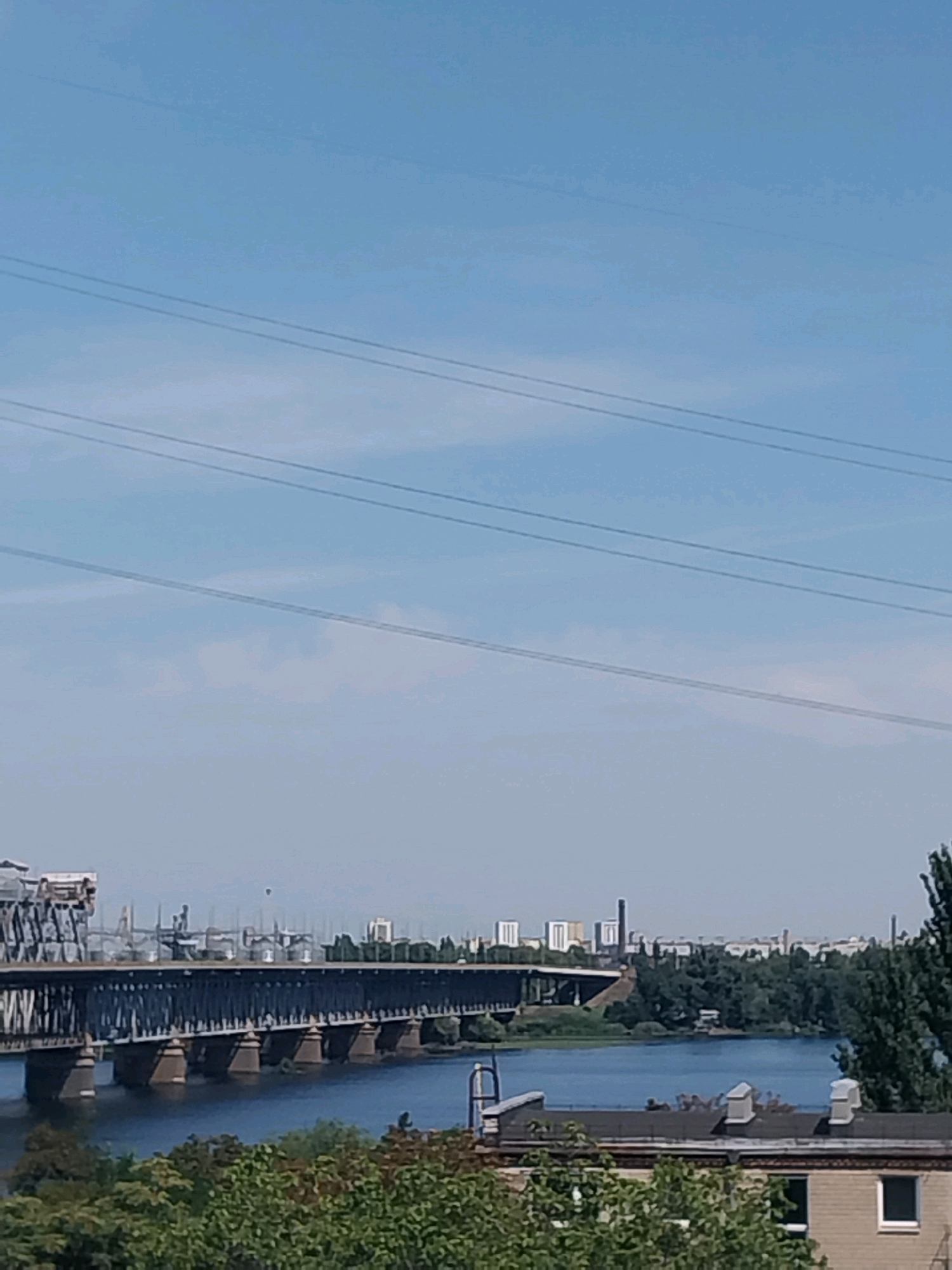 The image shows a wide river with a large bridge crossing it. The bridge is a steel truss structure with multiple piers supporting it. On the far bank, there are trees, buildings, and industrial structures. The sky is clear and blue with several power lines stretching across it. In the foreground, the tops of buildings and trees are visible. This scene depicts the Dnipro River in Dnipro, Ukraine, with the Amurskyi Bridge, a major railway bridge, prominently featured.