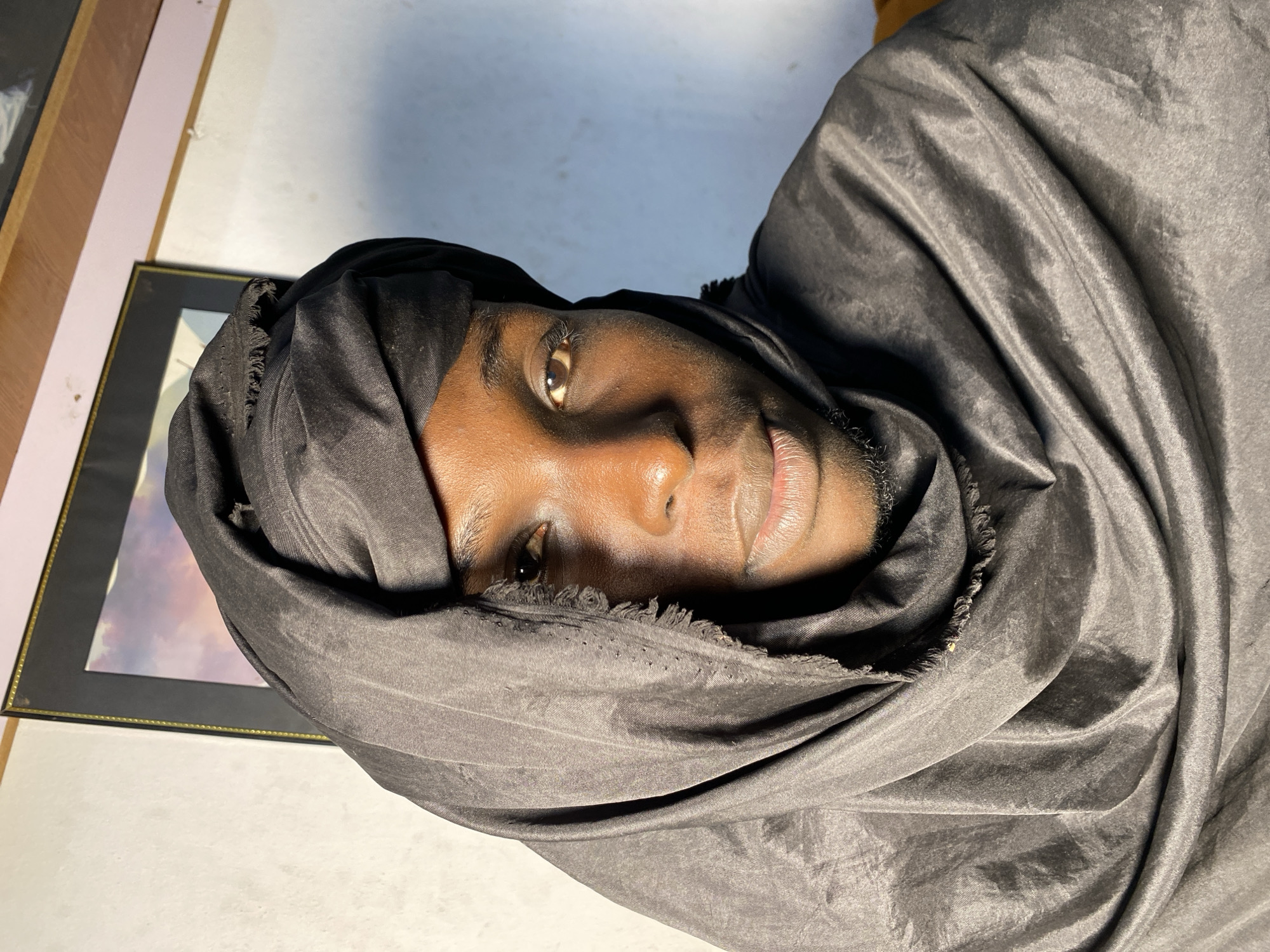 A medium close-up shot captures a young Black man from the chest up, wearing a black headwrap that covers most of his hair and forehead, leaving his face and neck exposed. The headwrap is made of a thin, flowing fabric, with a frayed edge visible on the left side of his head. His skin is a rich brown, with visible pores and subtle highlights from the lighting. He has short, dark eyebrows, brown eyes, a straight nose, and full lips. A faint shadow of stubble is visible on his chin.
The background is a plain white wall, with a framed picture hanging on it. The picture frame is black with a thin gold trim. The wall appears to be slightly textured and shows some minor discoloration. To the upper right of the frame, a wooden surface is visible, suggesting a shelf or ceiling trim.
The lighting appears to be from the right side, creating a shadow on the left side of the man's face and casting highlights on his forehead, cheekbones, and the fabric of his headwrap. The overall mood is contemplative and direct, with the subject making eye contact with the camera.
The image is believed to have been taken in Jos, Nigeria.