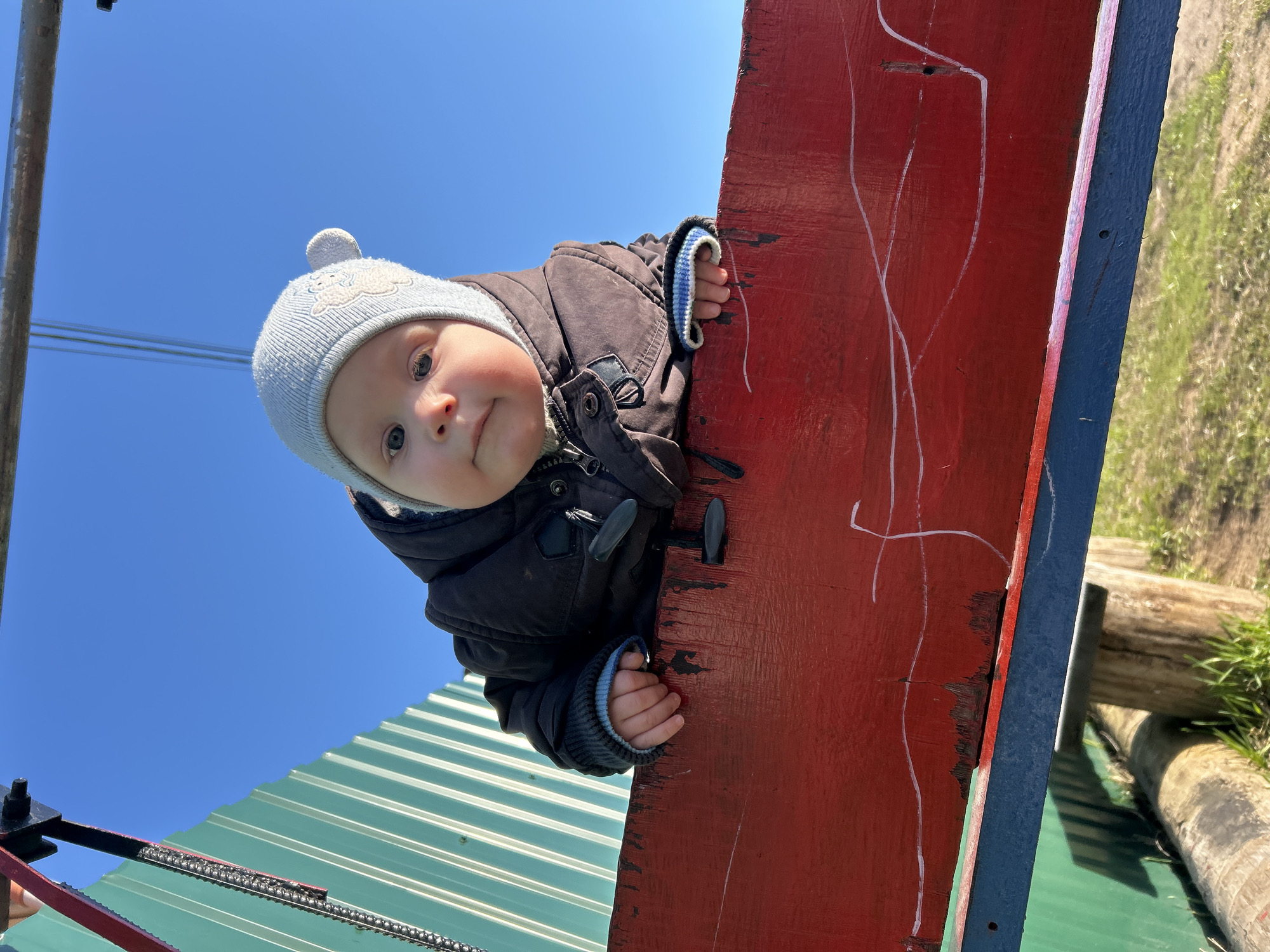 A baby wearing a light grey knit hat with a pom-pom and a dark brown winter coat is captured looking directly at the viewer. The baby is peering over a vibrant red wooden plank, holding onto its edge with both hands. The red plank, which features worn paint and faint white, squiggly markings, occupies a significant portion of the right side of the image.
In the background, a clear, bright blue sky fills the upper left area, with a metal pole or structure visible along the far left edge. Below the baby, parts of a corrugated green metal roof or surface are visible, alongside a black metal mechanism that includes a chain-like component. In the upper right corner, behind the red plank, a grassy area with rough wooden logs suggests ground level.
The scene takes place outdoors on a sunny day, indicated by the clear sky and bright lighting. The overall perspective is a low-angle shot, looking up at the baby. The location context for this scene is Chaikovsky, Russia. There is no visible text in the image.