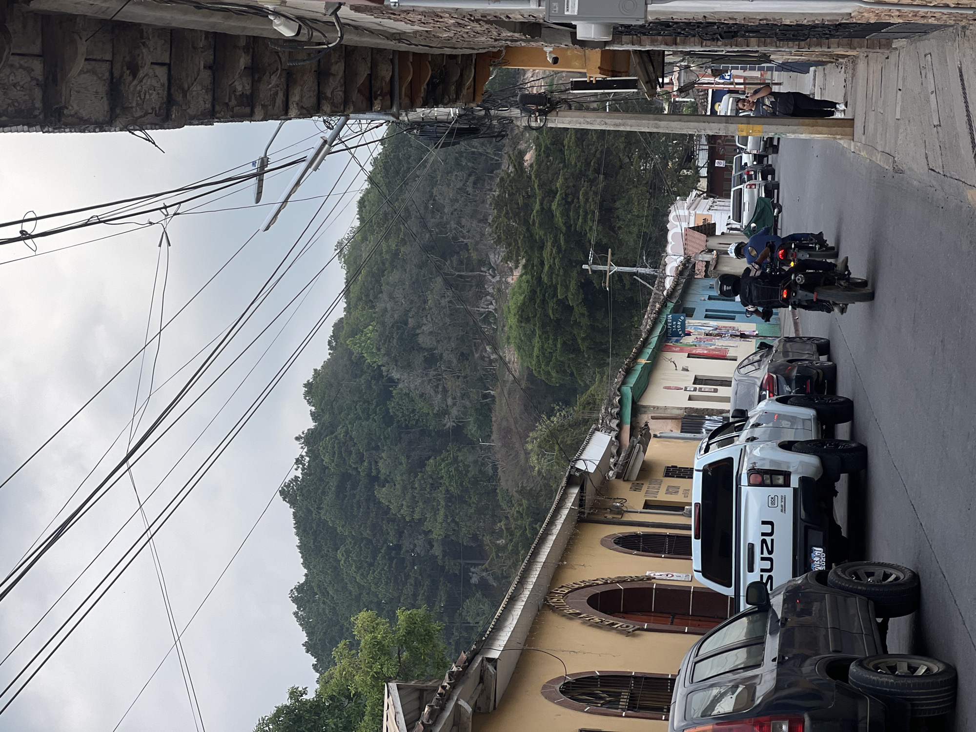A street in La Esperanza, Honduras is captured in this ground-level photograph. Two motorcyclists, one wearing a blue shirt and the other wearing a black shirt, are riding past parked cars on a street lined with buildings. A white Isuzu pickup truck and a dark SUV with spare tires piled on top are prominently parked on the right side of the road. In the distance, a person stands on the sidewalk, holding a phone up to their ear. The backdrop is dominated by a tree-covered mountain, with numerous power lines visible against the overcast sky. Architectural details of the buildings, including arched windows and stone facades, are observable along the sides of the road.