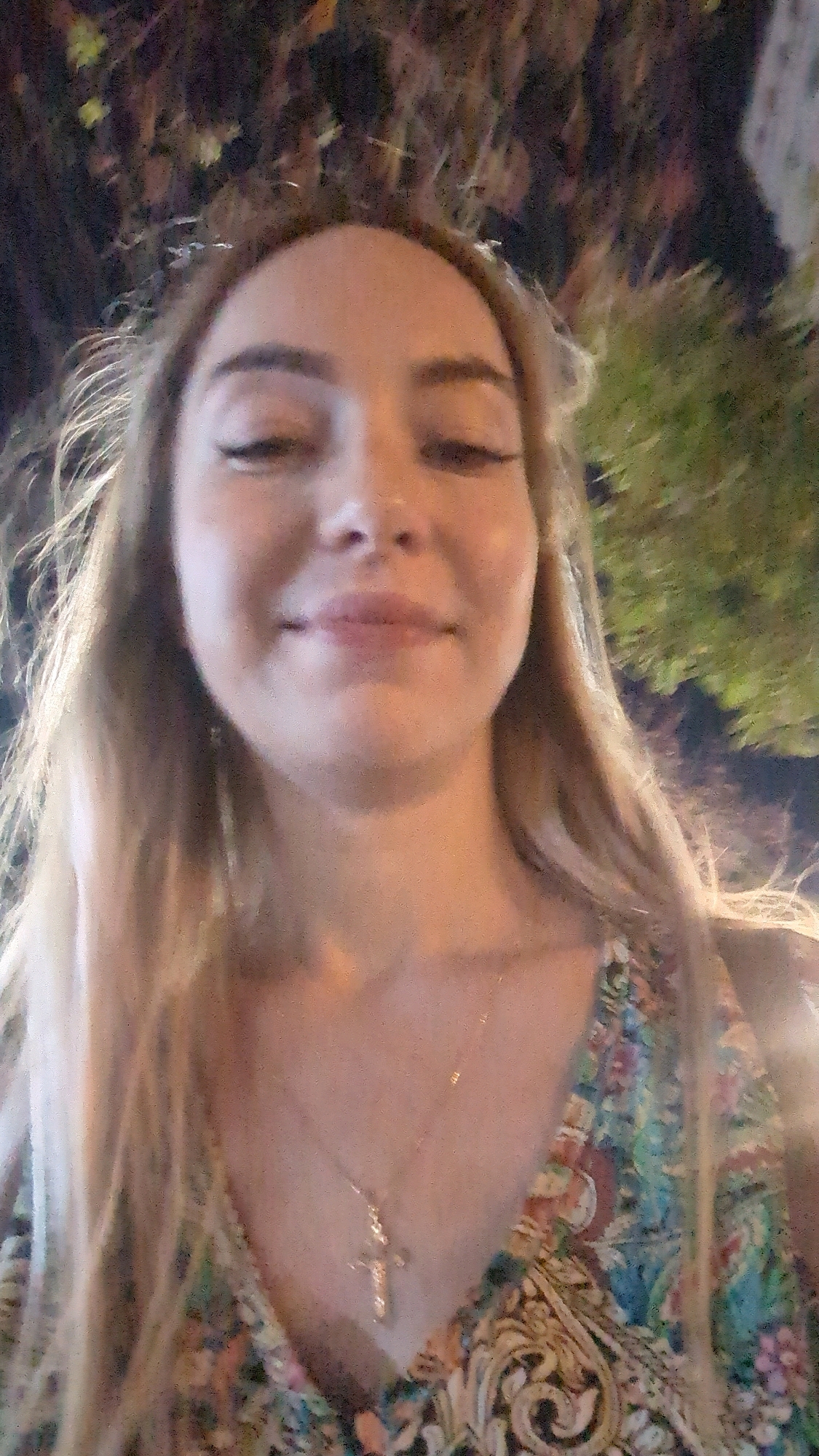 The image is a close-up selfie of a young woman with blonde hair, wearing a patterned dress and a cross necklace. She is looking down and smiling slightly. The background is blurred and appears to be outdoors, possibly at night, with foliage visible. There is no discernible information in the image that indicates the location is Chișinău, Moldova.