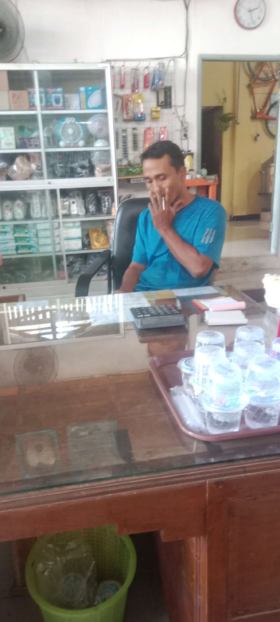 In Kras, Indonesia, a man is seen smoking a cigarette while sitting at a glass-topped counter in what appears to be a retail store. The man is wearing a blue t-shirt and is seated in a black office chair. Behind him, shelves are stocked with various items, including what look like fans, boxes, and packaged goods. A calculator and some papers are on the counter in front of him. To the right of the counter, a tray holds several small plastic cups of water and some wrapped straws. In the background, a doorway leads to another room where a bicycle is visible. A clock with red hands is mounted high on the wall. A green trash can is situated below the counter, containing plastic items. The overall environment suggests a commercial setting with merchandise and customer service elements.