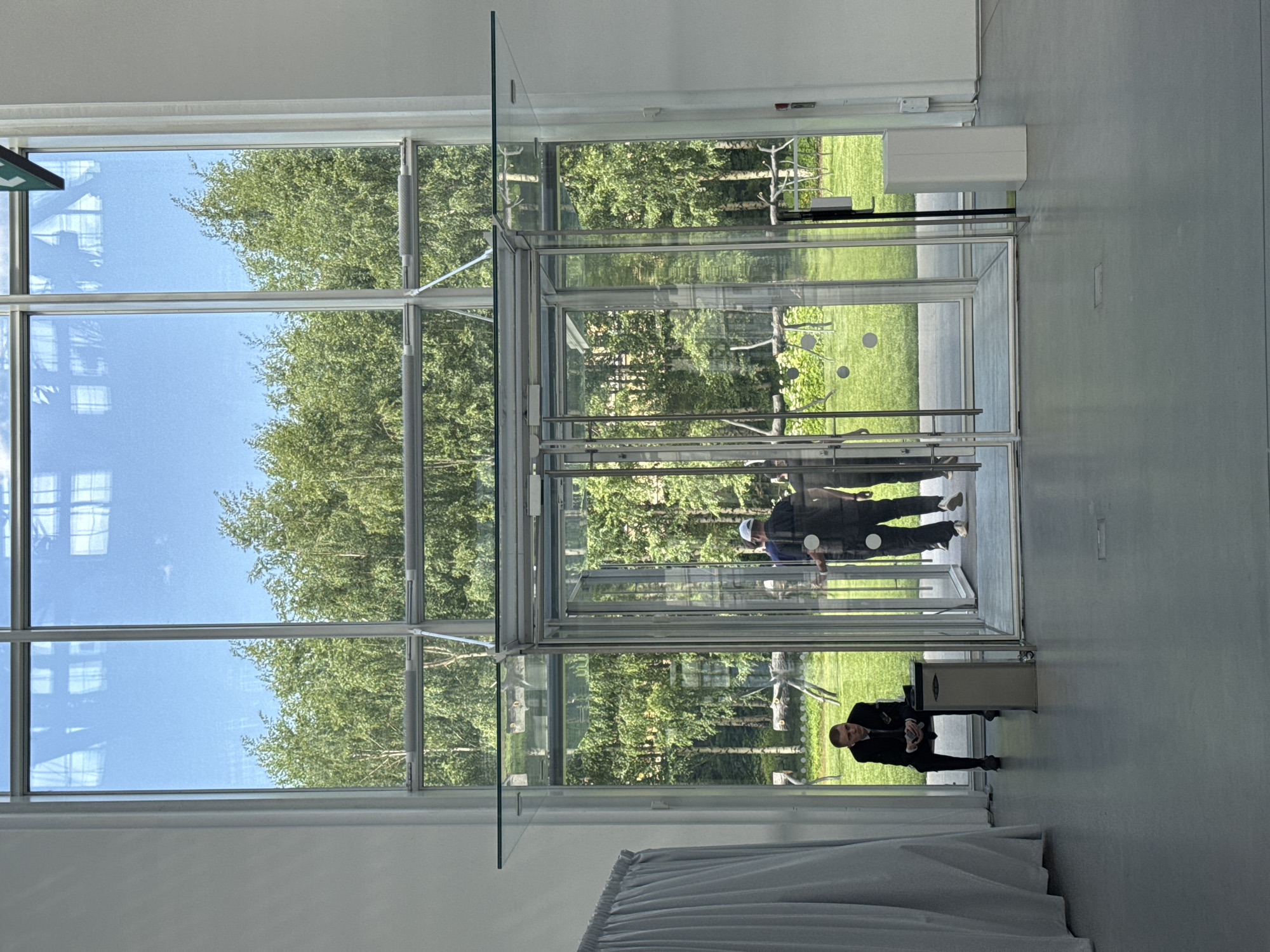 The image, oriented correctly, displays the interior of a modern building, likely in Moscow, Russia, characterized by extensive glass walls and doors that offer a clear view of the outside. The setting suggests a bright, sunny daytime, with a clear blue sky and lush green trees visible through the glass, possibly birches, creating a park-like environment.
In the foreground, a man in a dark suit with short hair is positioned near a silver rectangular bin or trash can. He appears to be standing or leaning, looking towards the viewer or slightly off-camera, potentially acting as a greeter, security, or an attendant at an entrance.
The building's interior features light-colored walls and flooring. A prominent glass structure, possibly a revolving door or an airlock-style entry system, dominates the central part of the image. Reflections of at least two other individuals are visible in these glass surfaces. One person, wearing a white cap and a dark top, is reflected within the glass entry system, suggesting movement into or out of the building.
The overall scene depicts a spacious and well-lit modern architectural space seamlessly connected to nature through its transparent design. There is no visible text in the image.