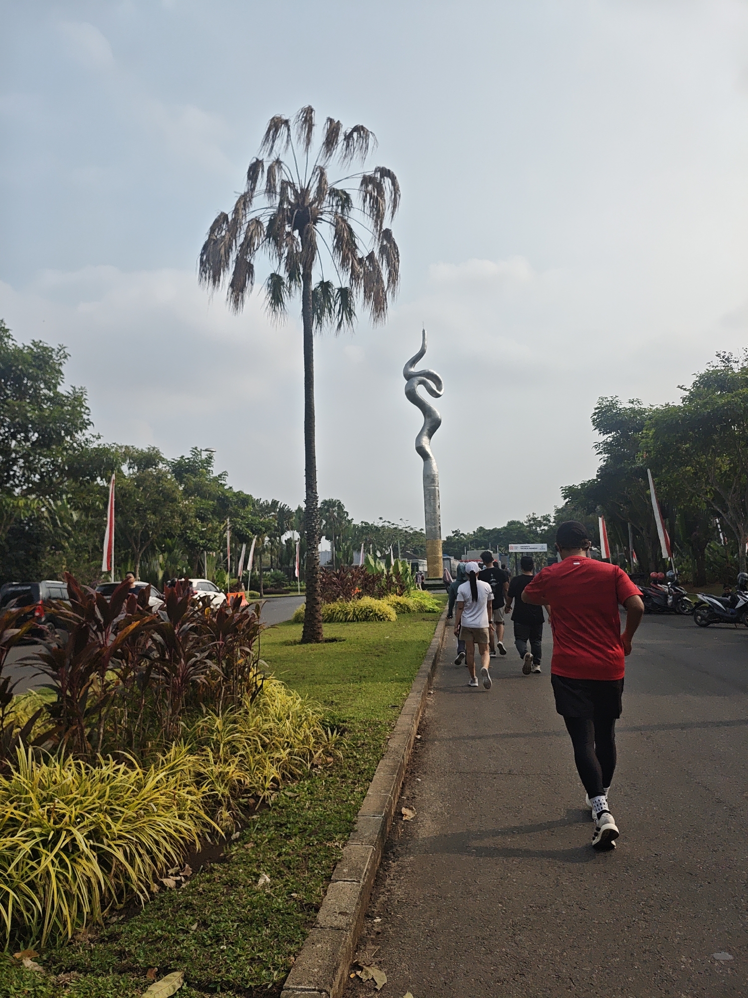 The image depicts a street scene in Bacin, Indonesia, with a group of people jogging on a paved road. In the foreground, a man in a red t-shirt and black shorts is running away from the camera. Behind him, several other individuals are also running or walking, including a person in a white t-shirt and khaki shorts. The road is bordered by a grassy area with ornamental plants, including a row of yellow, grass-like foliage and darker, broad-leafed plants. A tall, slender palm tree with dry fronds stands to the left of the road. In the background, a tall, abstract metallic sculpture, resembling a twisting ribbon or snake, is visible. Several cars are parked along the side of the road, and motorcycles are also present. Red and white flags are displayed on poles, suggesting a national holiday or event. The sky is overcast with a few clouds. The overall environment appears to be an outdoor recreational or park area within the city.