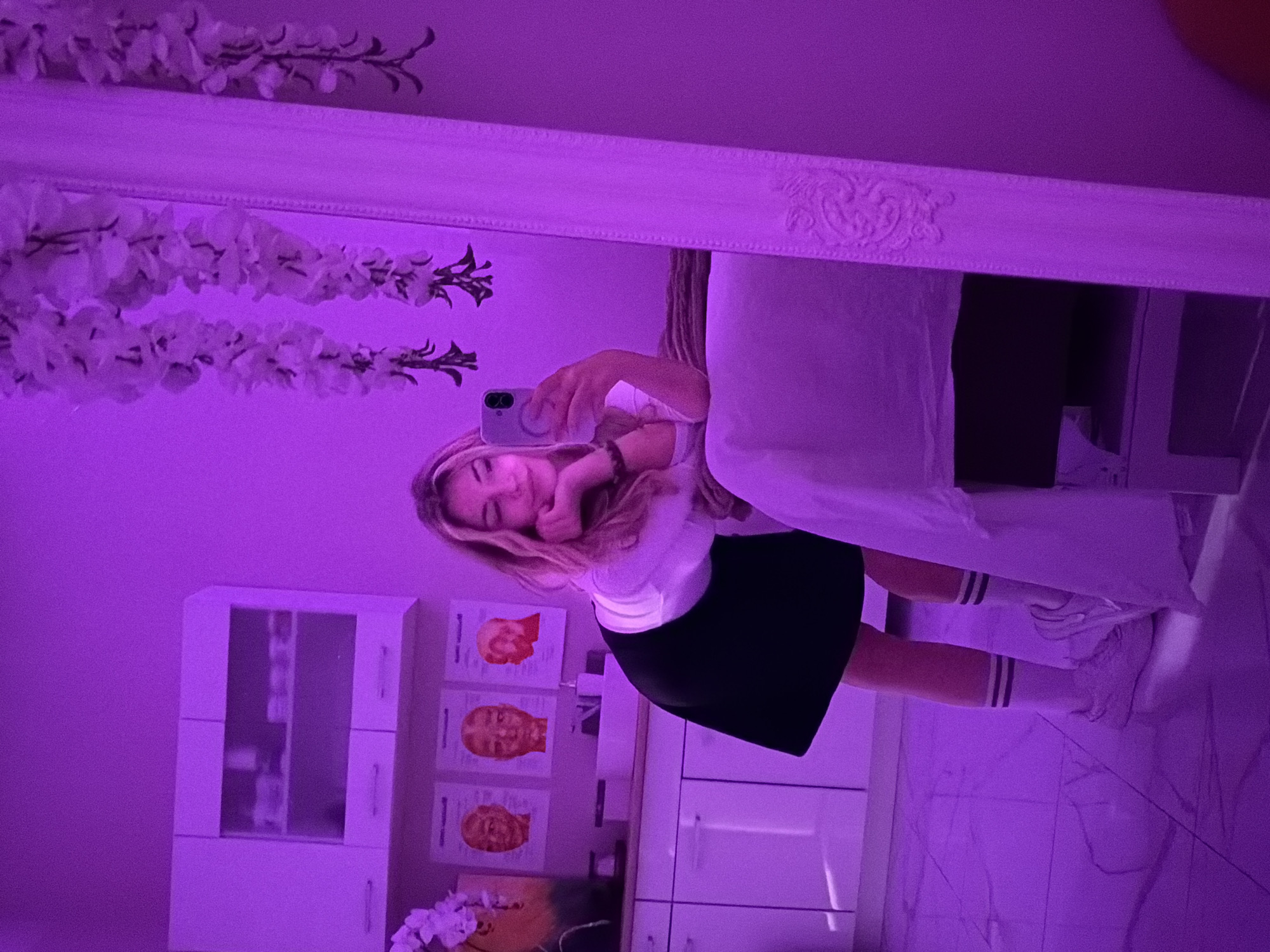 A young woman is captured taking a mirror selfie in an indoor setting, illuminated by strong purple ambient lighting. She has blonde hair and is wearing a white short-sleeved top, a black mini-skirt, white knee-high socks with two dark stripes, and white sneakers. She holds a light-colored smartphone in her right hand, extending it towards the mirror, and displays a gentle smile. A dark bracelet is visible on her left wrist.
The large mirror features an ornate white frame. Along the top edge of the mirror, several strands of white artificial flowers are arranged. The room's interior, reflected in the mirror, shows a white cabinet with glass doors on the upper section and solid doors below, positioned to the left. On the wall above the cabinet, three framed posters depict red silhouettes of human heads, possibly illustrating anatomical charts or cosmetic procedure diagrams. Below these, another poster with a yellow/orange facial illustration is partially visible. To the right of the woman, a white surface, likely a bed or a treatment table, is covered with white linens. The floor appears light-colored, possibly tiled. The pervasive purple light creates a distinct, modern, or stylized atmosphere for the scene, which hints at a beauty salon, spa, or clinic setting due to the displayed posters and treatment-like furniture. No distinct text is legible on the posters or elsewhere in the image.