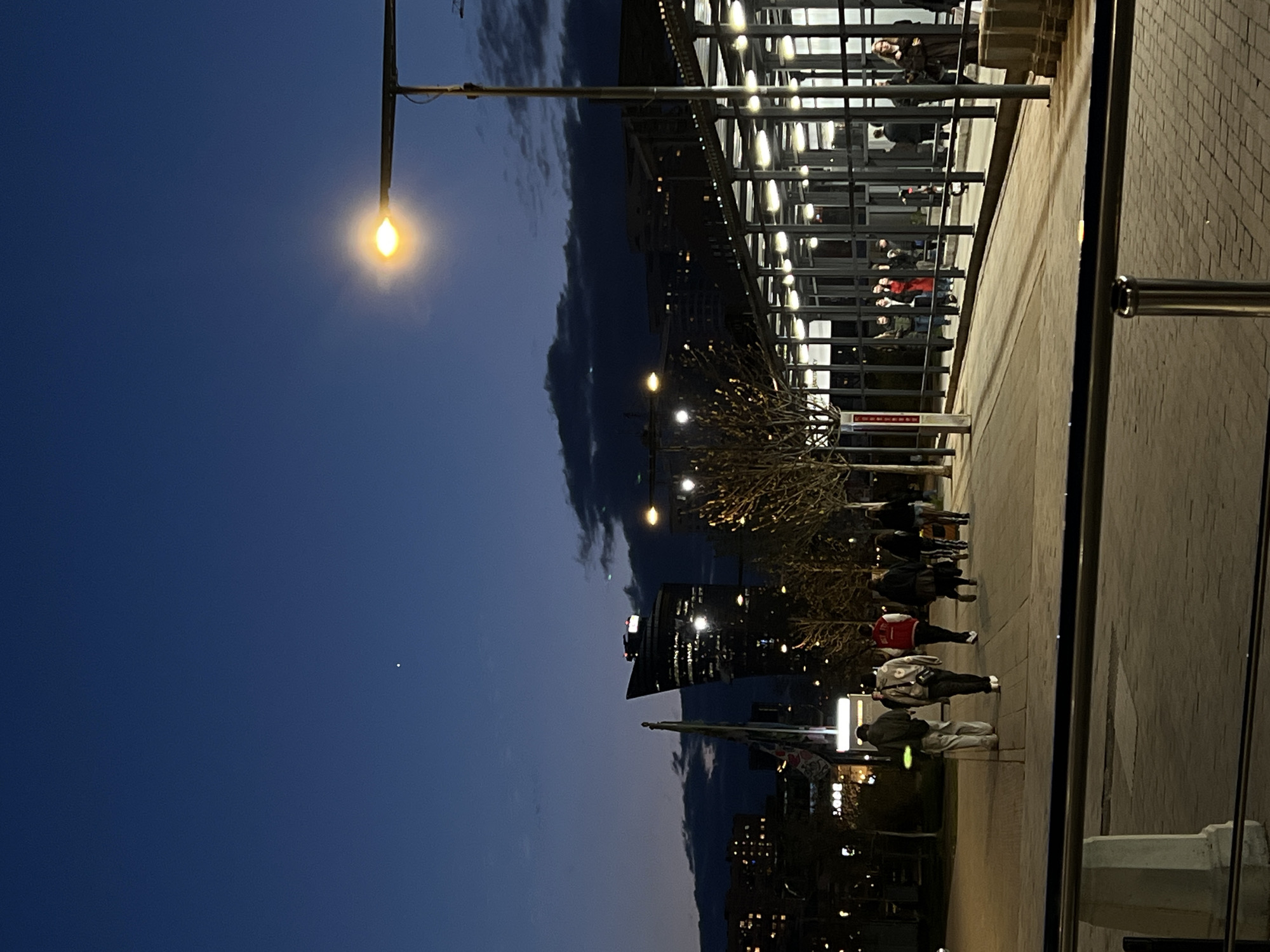 The image shows an evening scene in Khimki, Russia. People are walking along a paved walkway next to a modern building with glass walls and a steel frame. There is a streetlight casting a yellow glow. A tree with bare branches stands next to the walkway. In the background, there are other buildings. The sky is a gradient of blue and dark grey, with some clouds visible. A flag is planted at the base of the buildings.