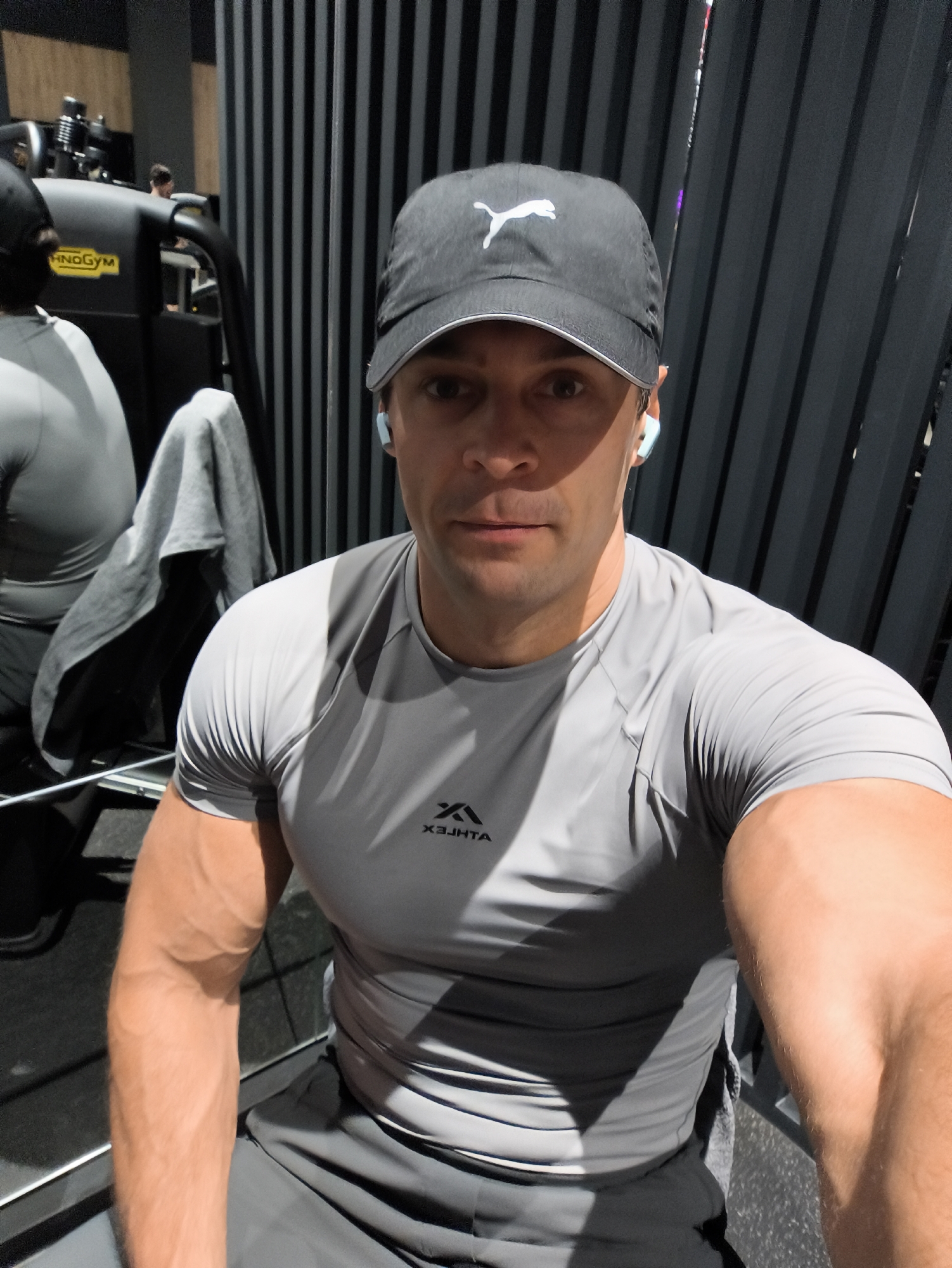 A man is taking a selfie in a gym. He is wearing a grey t-shirt, grey shorts, a black baseball cap with a white Puma logo, and light blue earbuds. The man has muscular arms and is looking at the camera. In the background, there is gym equipment, including a weight machine with the "HOGYM" logo visible, and a mirror reflecting another person. The setting appears to be indoors with vertical black slats on the wall behind the man. The lighting suggests it is daytime. There is a grey towel draped over a piece of gym equipment. The t-shirt has a small, upside-down logo on the chest that reads "XEHJTA".