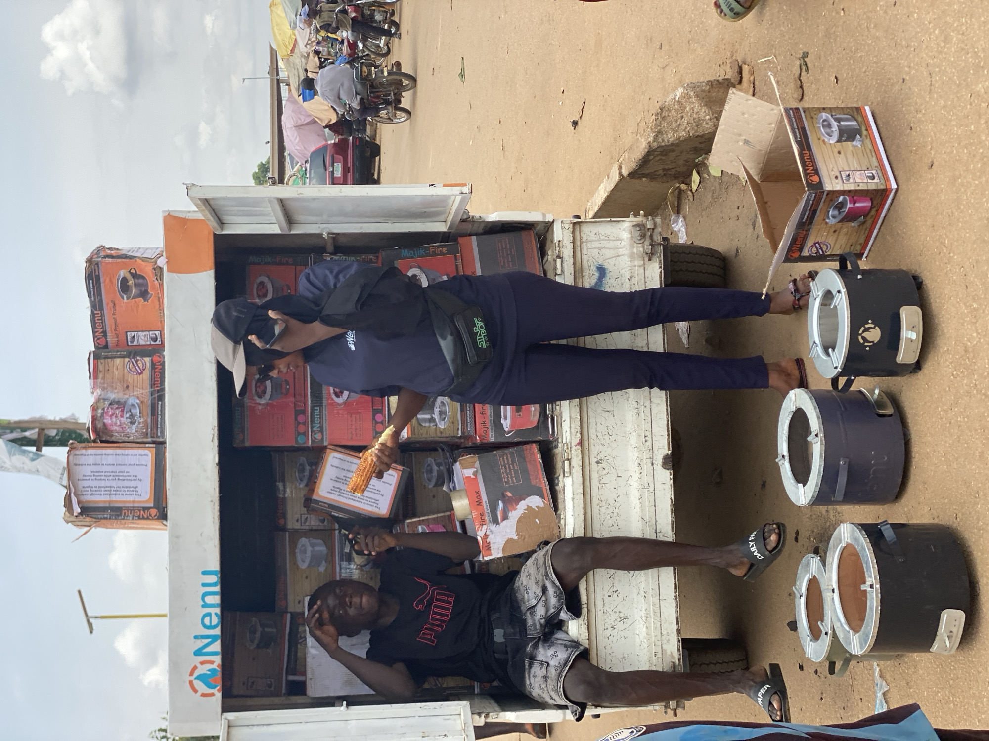 This image captures a vibrant outdoor market scene, likely in Kafanchan B, Nigeria, under bright daylight with a partially cloudy sky. The focal point is a white pickup truck, its back open and filled with numerous cardboard boxes, suggesting it's being used as a mobile shop.
Two males are prominently featured. One man, dressed in a navy blue top, dark trousers, and a baseball cap, stands inside the truck bed. He is holding a piece of roasted corn and wears a black fanny pack with "STBBY" printed on it, and his sandals bear the text "DAILY PAPER." The second man sits on the edge of the truck bed, wearing a black t-shirt with "PUMA" clearly visible and patterned shorts, appearing relaxed.
The truck's cargo consists of boxes branded "Nenu" and "Majik-Fire," with product images indicating cooking stoves or similar kitchen appliances. Several unpacked, dark-colored metal charcoal stoves are displayed on the dusty ground next to the truck, along with an open "Nenu" branded box. In the background, a busy street or market area is visible, with motorcycles, a red car, and makeshift stalls covered by tarpaulins, hinting at a lively commercial environment. The scene depicts the sale or distribution of these household items from the truck.