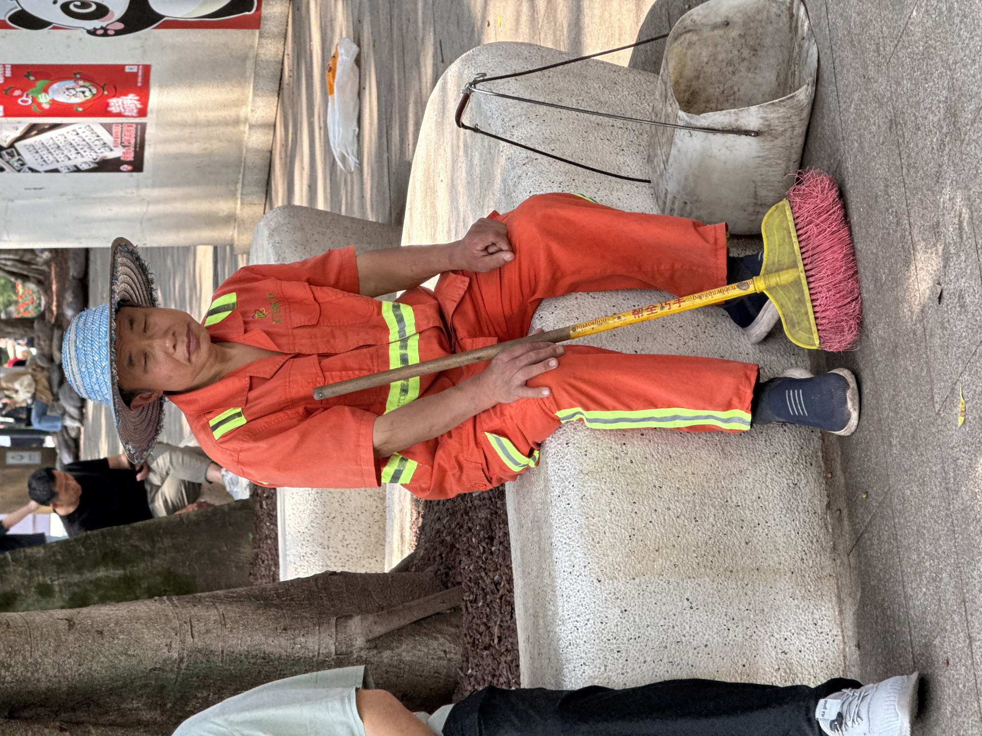 A man wearing an orange jumpsuit and a blue straw hat sits on a light-colored stone structure, holding a yellow broom with pink bristles. He is also wearing navy blue sneakers with white soles. To his right is a white bucket. The location is Jiulongpo District, China, indicated by the writing on the broom. There are other people in the background. A poster can also be seen on the wall behind the man.