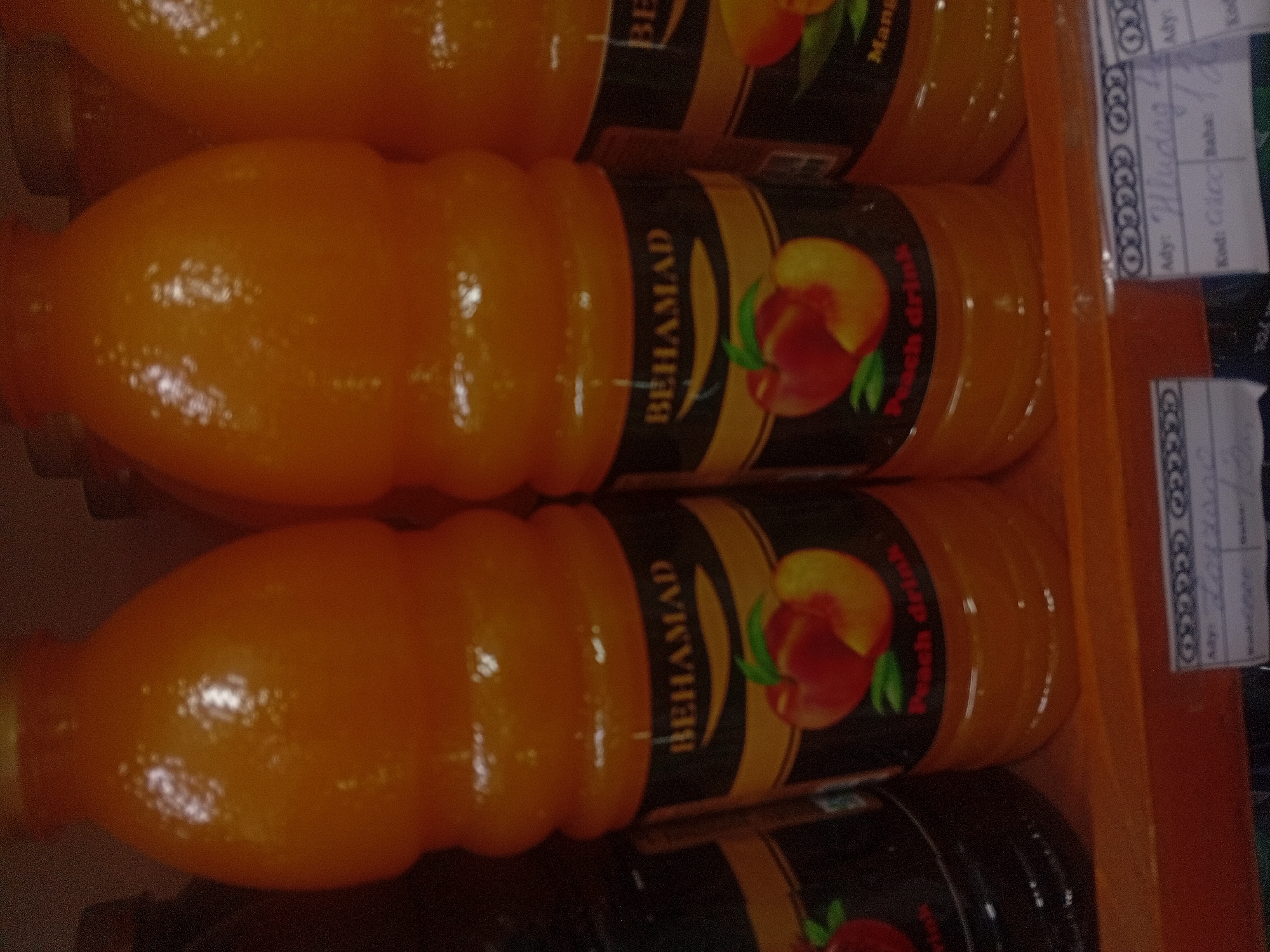 The image shows a shelf of beverages, likely in a store or market in Ashgabat, Turkmenistan. The focus is on several large, orange-colored plastic bottles filled with a liquid that appears to be juice. The bottles have labels with branding, including the word "BEHAMAD" and images of peaches. Some labels also indicate "Peach drink." In the foreground, there are price tags or labels with text in what appears to be Cyrillic script, suggesting a regional origin. The lighting is somewhat dim, and the overall image quality is low, with some blurriness.