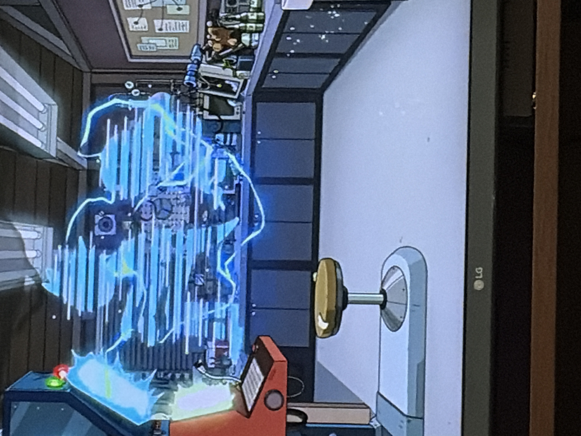 The image depicts an animated scene inside a laboratory. A large, intricate device emitting blue energy and light is the central focus, positioned on a workbench. Various laboratory equipment, including monitors, bottles, and what appears to be a teddy bear, are visible on the workbench. In the foreground, a stool with a yellow seat stands on a light-colored floor. The room has dark cabinets lining the walls and fluorescent lighting fixtures on the ceiling. The scene suggests an ongoing experiment or technological operation within the laboratory setting. The location is identified as Unknown, Unknown.