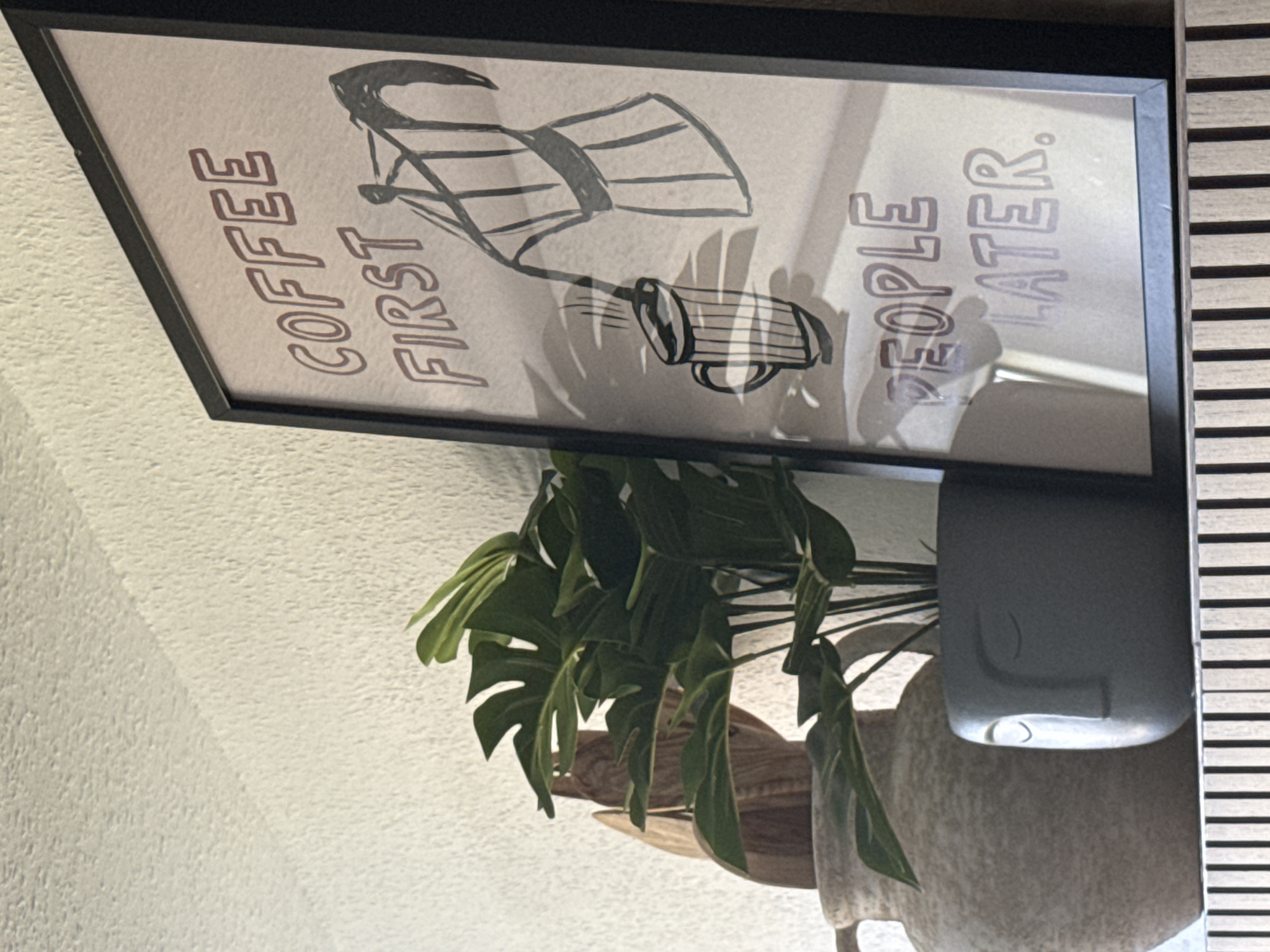 The image shows a shelf with various decorative items, including a framed poster that reads "COFFEE FIRST PEOPLE LATER." The poster features a hand-drawn illustration of a moka pot pouring coffee into a mug. Next to the poster is a potted plant with large green leaves, placed in a planter shaped like a face. Behind the plant is a large, textured vase. The shelf itself has a striped wooden surface. The background is a textured white wall. The scene appears to be an indoor setting, possibly a home or a cafe. There is no information available to determine the specific location of this scene in Chemnitz, Germany.