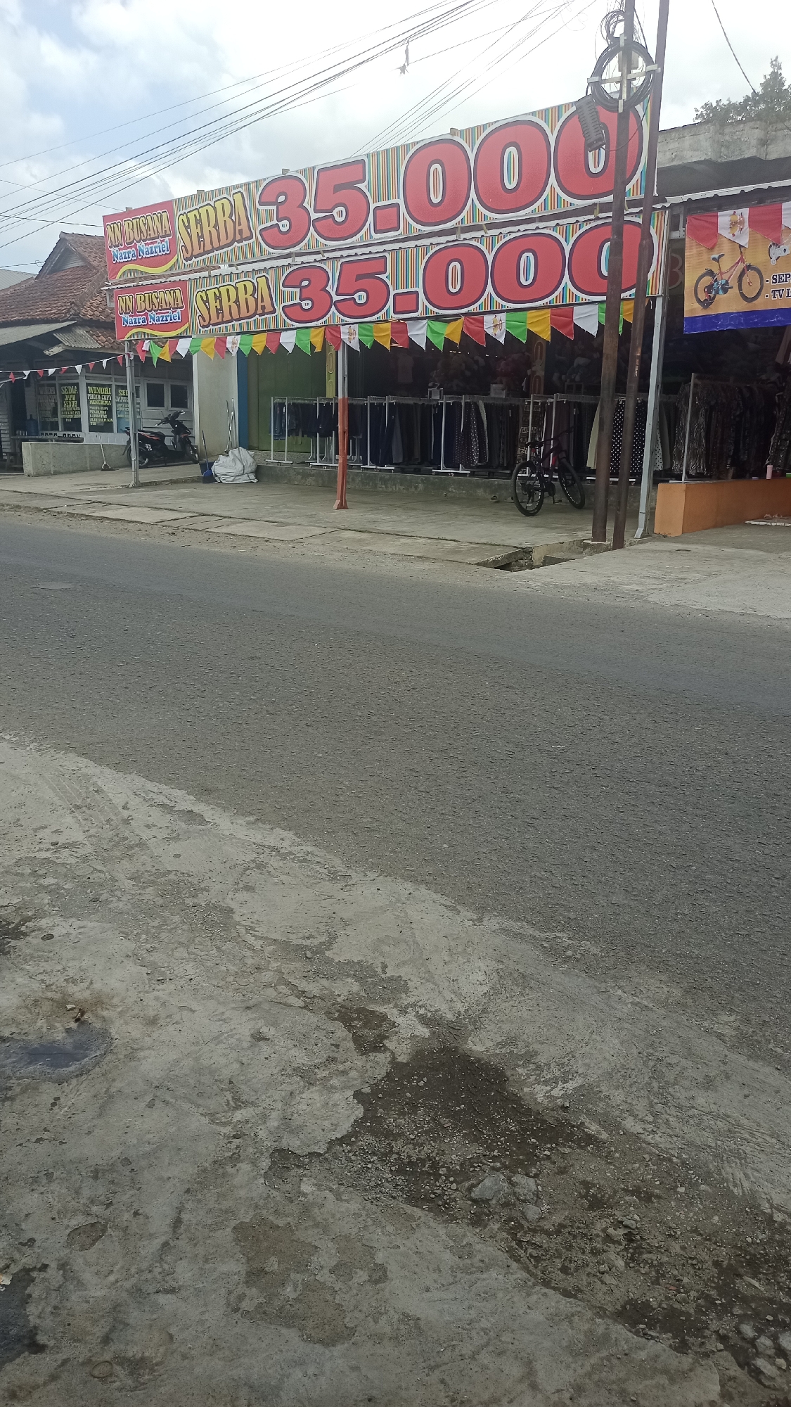 The image depicts a street scene in Cikalong, Indonesia. In the foreground, a paved road with some debris and dark patches is visible. Across the road, a store with a large banner advertising "SERBA 35.000" (meaning "Everything 35,000") is prominent. The banner features large red numbers and is decorated with colorful pennants. The store appears to be selling clothing, with racks of garments displayed outside. A motorcycle is parked near the store, and a bicycle is also visible. The background shows a building with a traditional Indonesian roof. The sky is overcast, and overhead wires are present.