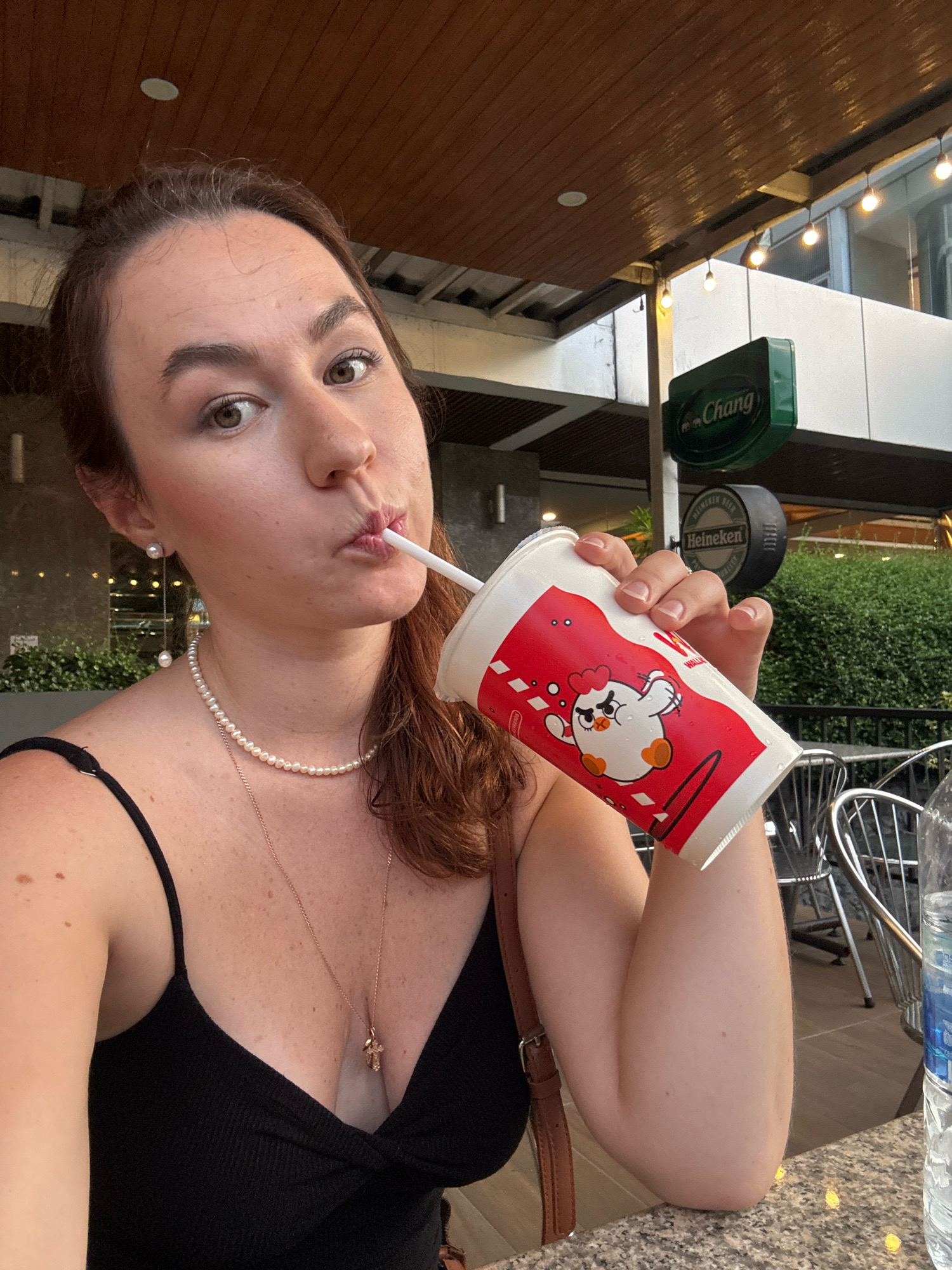 A young Caucasian woman with brown hair is shown in a close-up shot, drinking from a disposable cup with a red and white chicken graphic. She is wearing a black spaghetti strap top, a pearl necklace, and a gold cross pendant. Her left arm is raised, holding the cup to her lips with a straw. The background suggests an outdoor seating area, possibly a restaurant or cafe, with a wooden ceiling, modern light fixtures, and signage for "Chang" and "Heineken." The setting appears to be in Bangkok, Thailand, given the prominent "Chang" beer sign, a popular Thai brand.