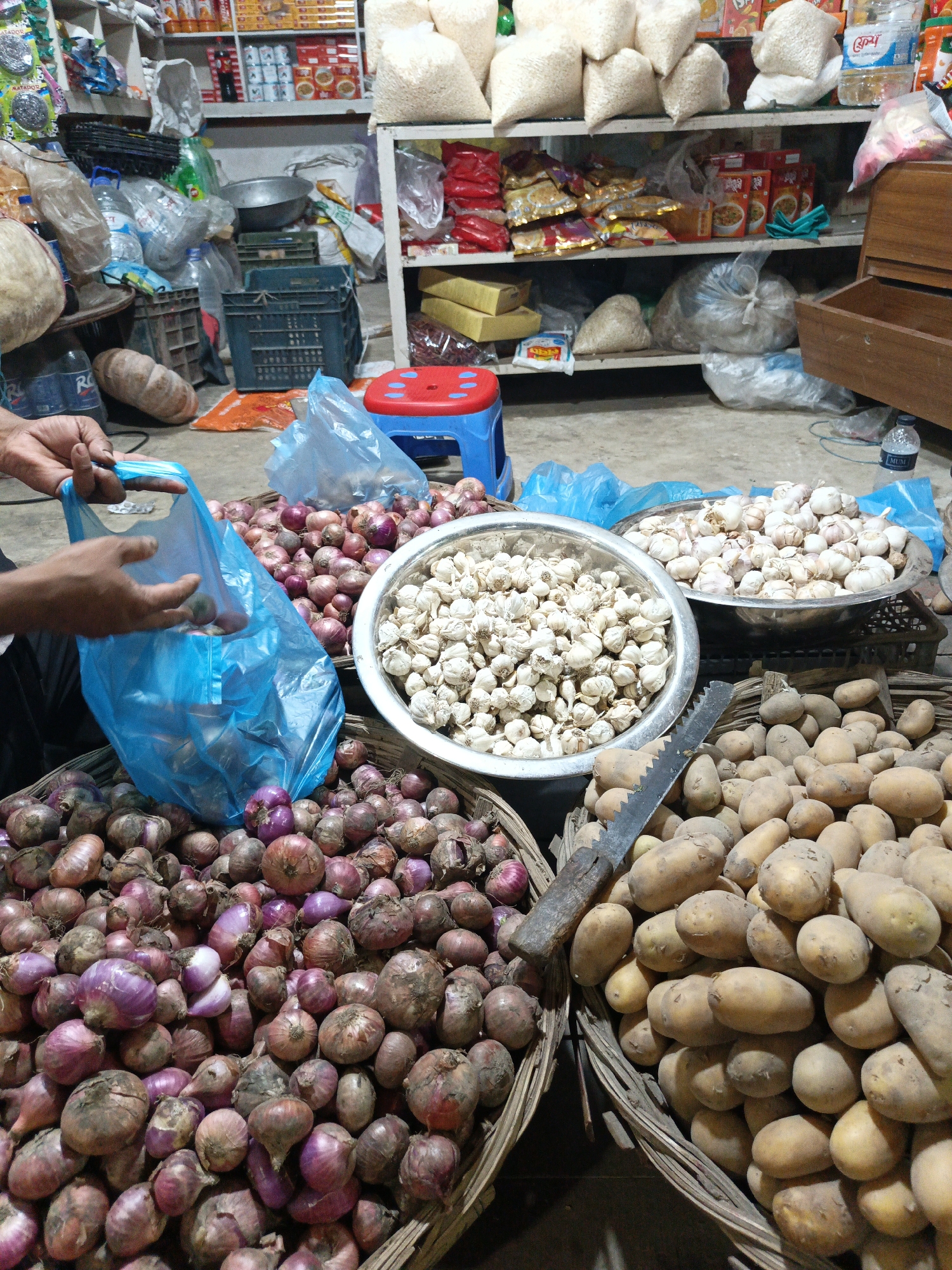 The image shows a vegetable stall in Dhaka, Bangladesh. There are several baskets and bowls filled with vegetables. In the foreground, there is a basket full of red onions and another basket full of potatoes. Two silver bowls are filled with garlic cloves. A serrated knife sits on top of the potatoes. A person's hands are visible holding a blue plastic bag over one of the onion baskets, likely filling it with onions. In the background, there's a shelf stocked with various packaged goods, bags of grains, and other food items, suggesting a small grocery store setting. A blue and red plastic stool is visible. The scene appears to be inside a shop or market area.