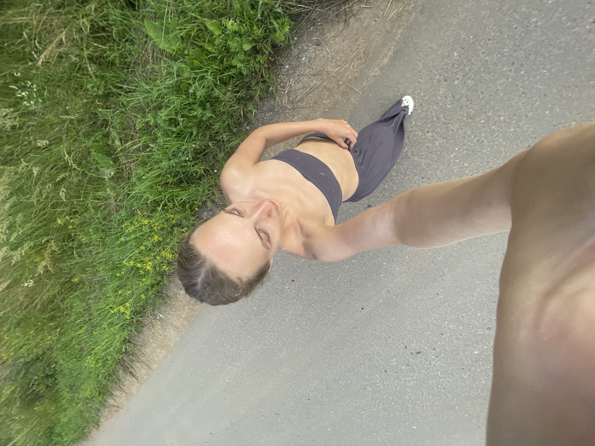 A woman is captured in a high-angle selfie, standing on an asphalt path bordered by lush green grass. She is wearing a dark gray sports bra and matching loose-fitting pants, with white athletic shoes on her feet. The woman is looking down at herself, with her right hand touching her stomach. Her hair is pulled back, and her expression appears contemplative. The scene suggests an outdoor activity, possibly a workout or a walk, given her attire and the setting. The lighting indicates it is daytime, and the greenery suggests a temperate season, likely spring or summer. The background reveals more of the unpaved verge and the textured asphalt. The image was taken in Подшивалово, Russia.