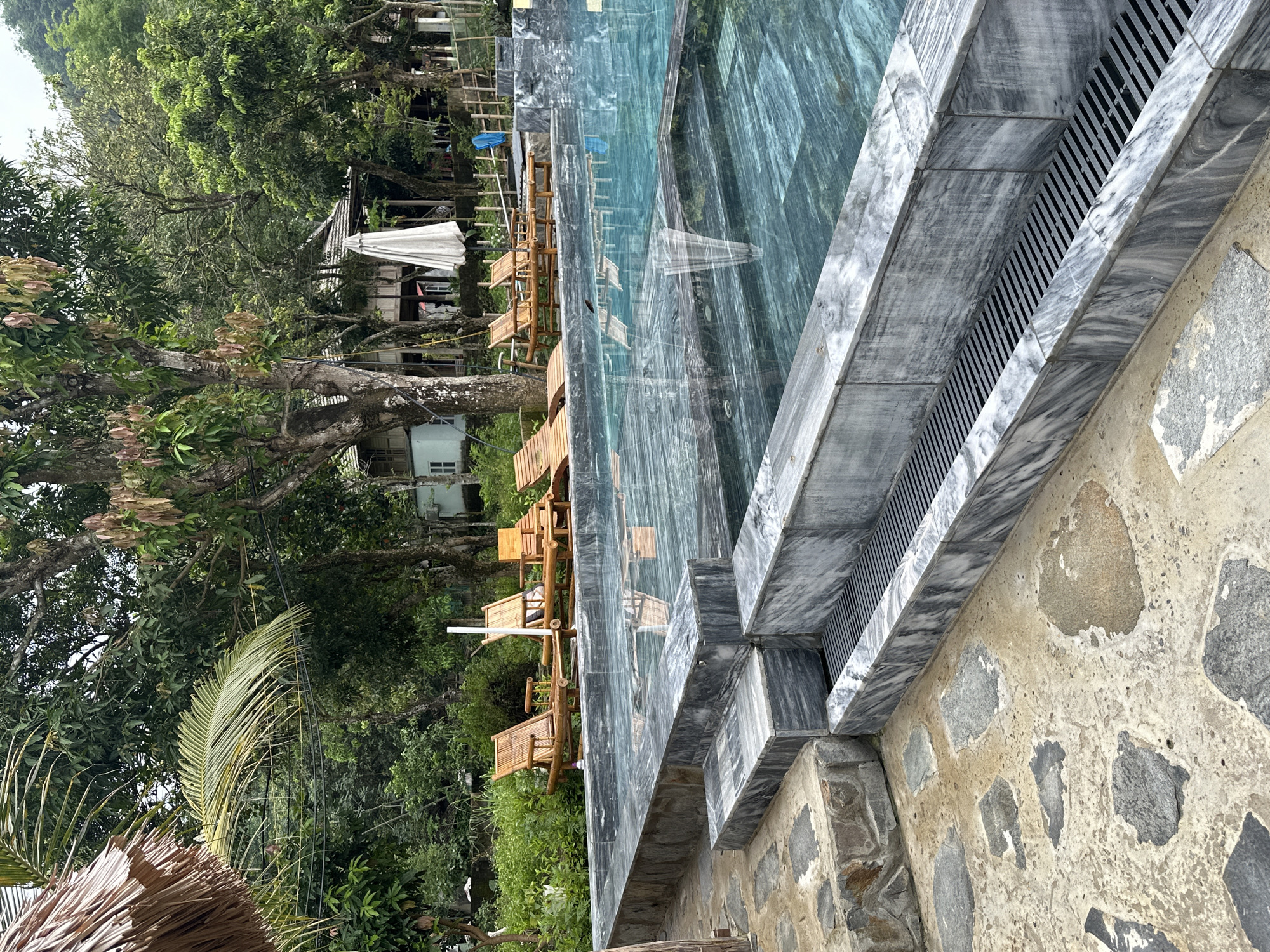A resort pool area is depicted in Xóm Khán, Vietnam. The pool has a marble surround and a tiled bottom that appears to be made of green marble. Several lounge chairs made of bamboo are positioned near the pool. In the background, trees and a building with a thatched roof are visible. A white umbrella is also present. The ground around the pool is paved with stone. The image appears to have been taken on an overcast day. There are no people or animals visible in the image.