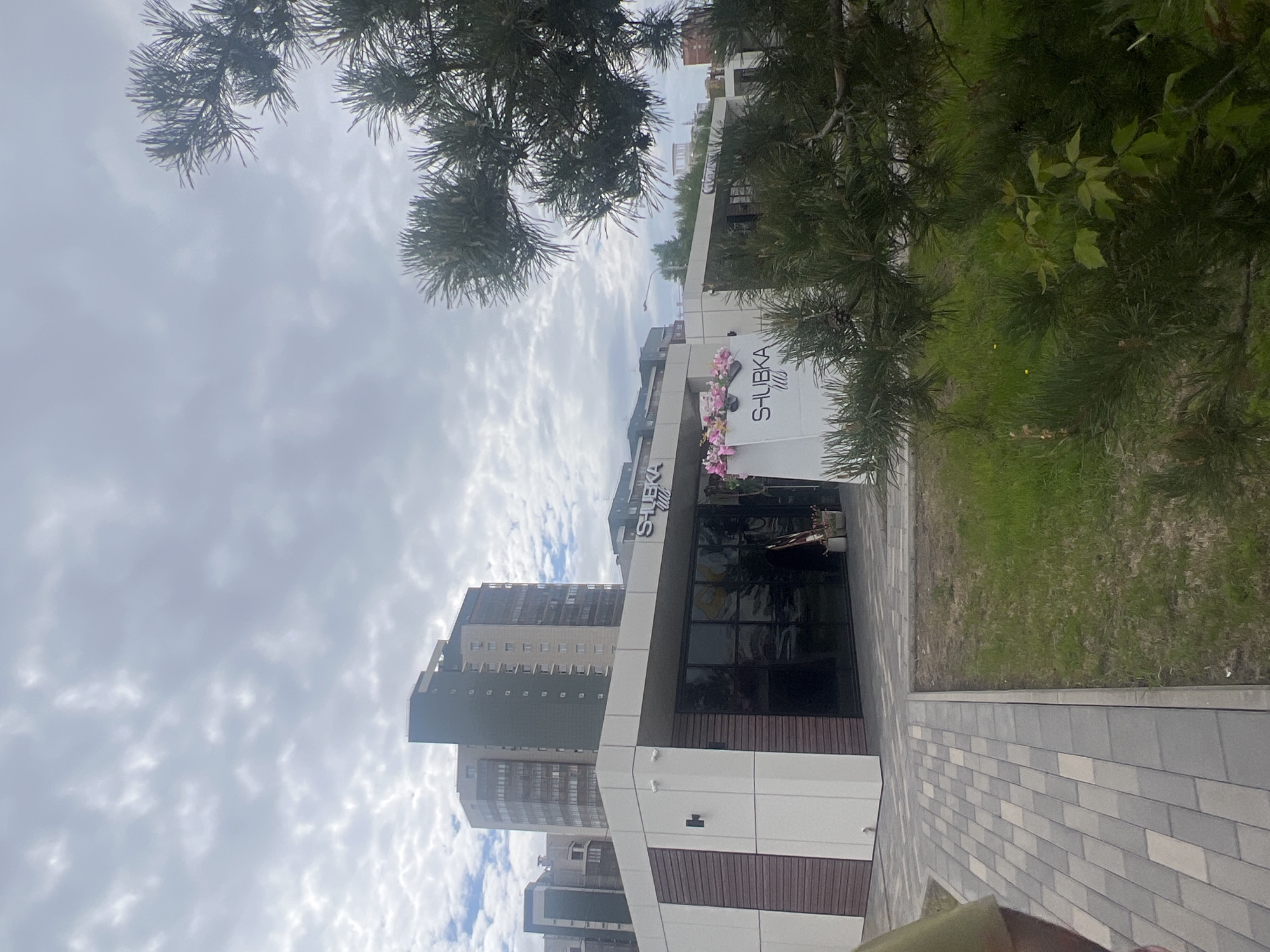The image shows a building with the signage "SHUBKA Club" located in Krasnoyarsk, Russia. The building has a modern design with white and brown paneling, and large glass windows. A high-rise building is visible in the background. The sky is cloudy. The ground is paved with gray bricks, and there is a grassy area to the side. Trees are visible framing the top of the image.
