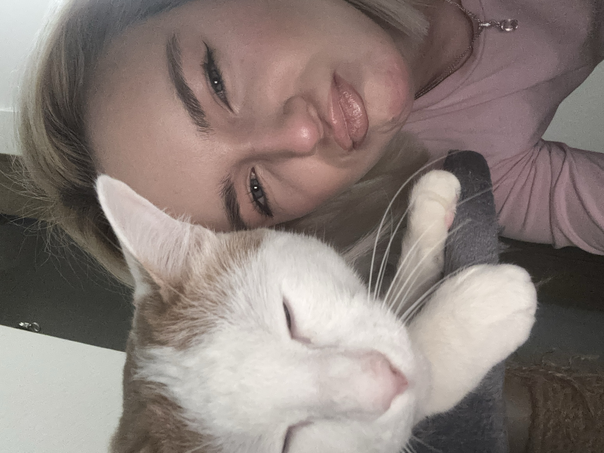 A selfie-style photo shows a woman and a cat. The woman, facing the camera at an angle, has light skin, light blonde hair, dark eyeliner, and pink lipstick. She is wearing a pink top and a necklace. The cat, predominantly white with patches of orange, is in the foreground. It is resting its paw on a gray, textured surface, possibly a scratching post or piece of furniture. The background is blurred and indistinct, suggesting an indoor setting. The location in Vsevolozhsk, Russia cannot be determined from the image.