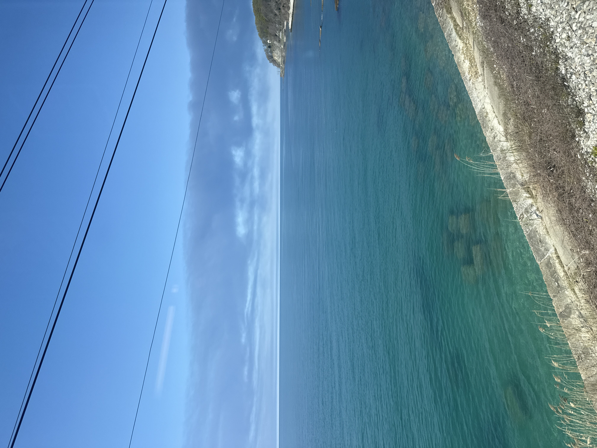 The image shows a view of a body of water, likely a lake or sea, with a rocky shoreline to the right. The water is clear and turquoise in color. In the distance, a rocky cliff or landmass is visible. To the left, the sky is blue with some wispy clouds. Three dark lines, likely power lines or cables, are visible against the sky. The image appears to have been taken from an elevated position, possibly a bridge or cliff overlooking the water. There is vegetation along the shoreline, including grasses and shrubs. The location is not specified.
