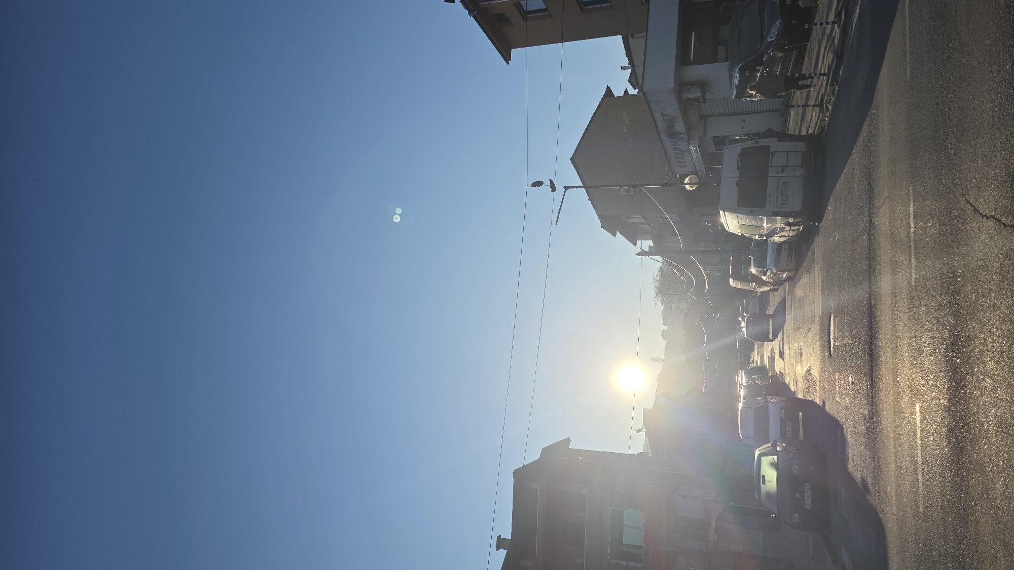 The image depicts a street scene in Blagoevgrad, Bulgaria, taken during a sunny day. The perspective is from street level, tilted 90 degrees. The sun is positioned in the distance, emitting a strong glare. On the right side of the image, there is a street with several parked vehicles, including vans and cars. A few individuals are walking on the sidewalk. Buildings of varying heights line the street, and power lines stretch across the sky. The clear blue sky occupies the left portion of the image, with two noticeable lens flares.