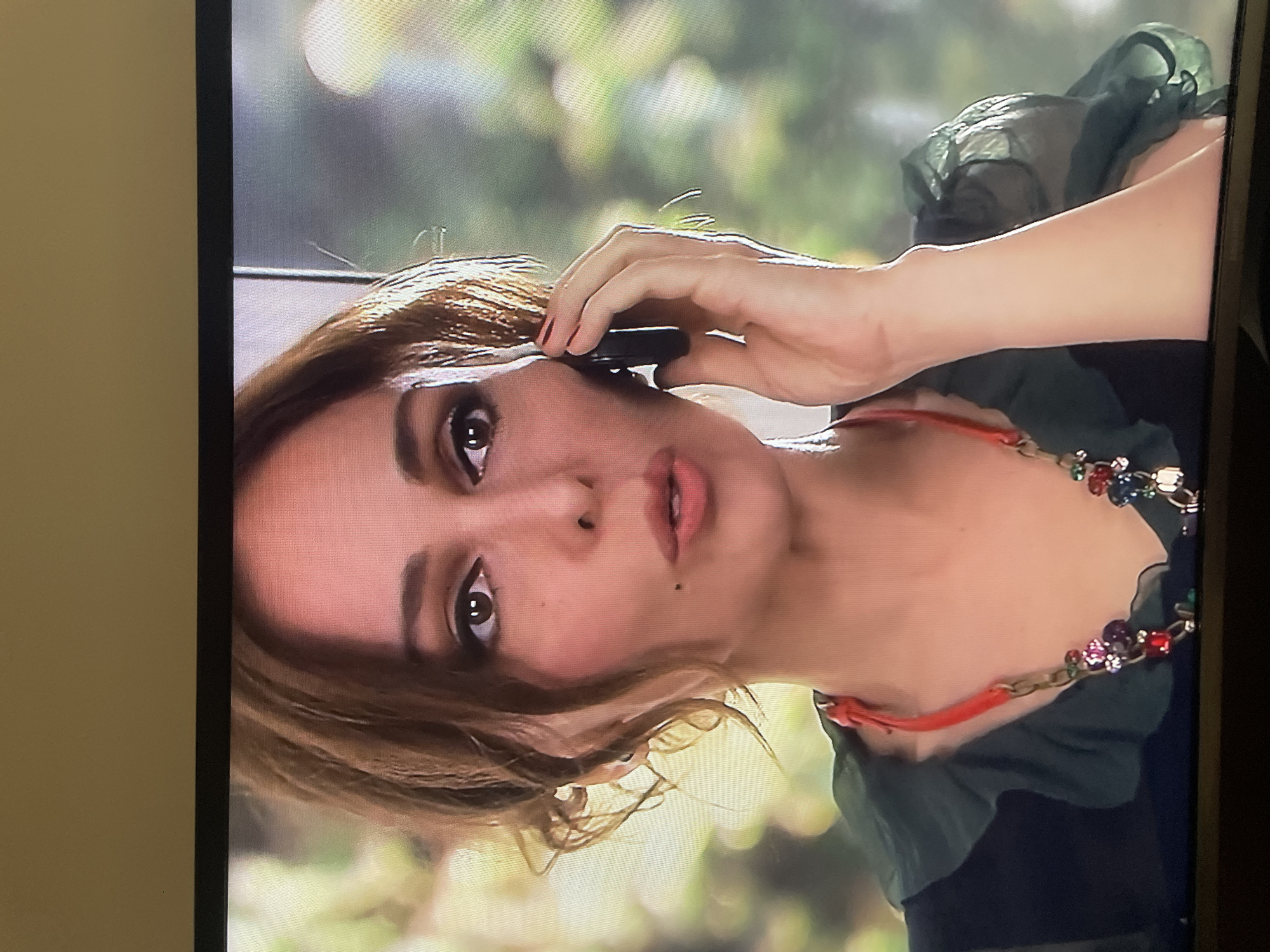 The media file shows a close-up of a woman speaking on a cell phone. She has brown hair styled loosely, with some strands framing her face. Her makeup is noticeable, with dark eyeliner and mascara, and she has a mole on her right cheek. She is wearing a necklace with colorful gemstones. Her expression is somewhat intense, suggesting she is engaged in a serious conversation. The background is blurred, but appears to be an outdoor setting with greenery and sunlight, possibly indicating daytime. The overall composition is a portrait-style shot focusing on the woman's face and upper body.