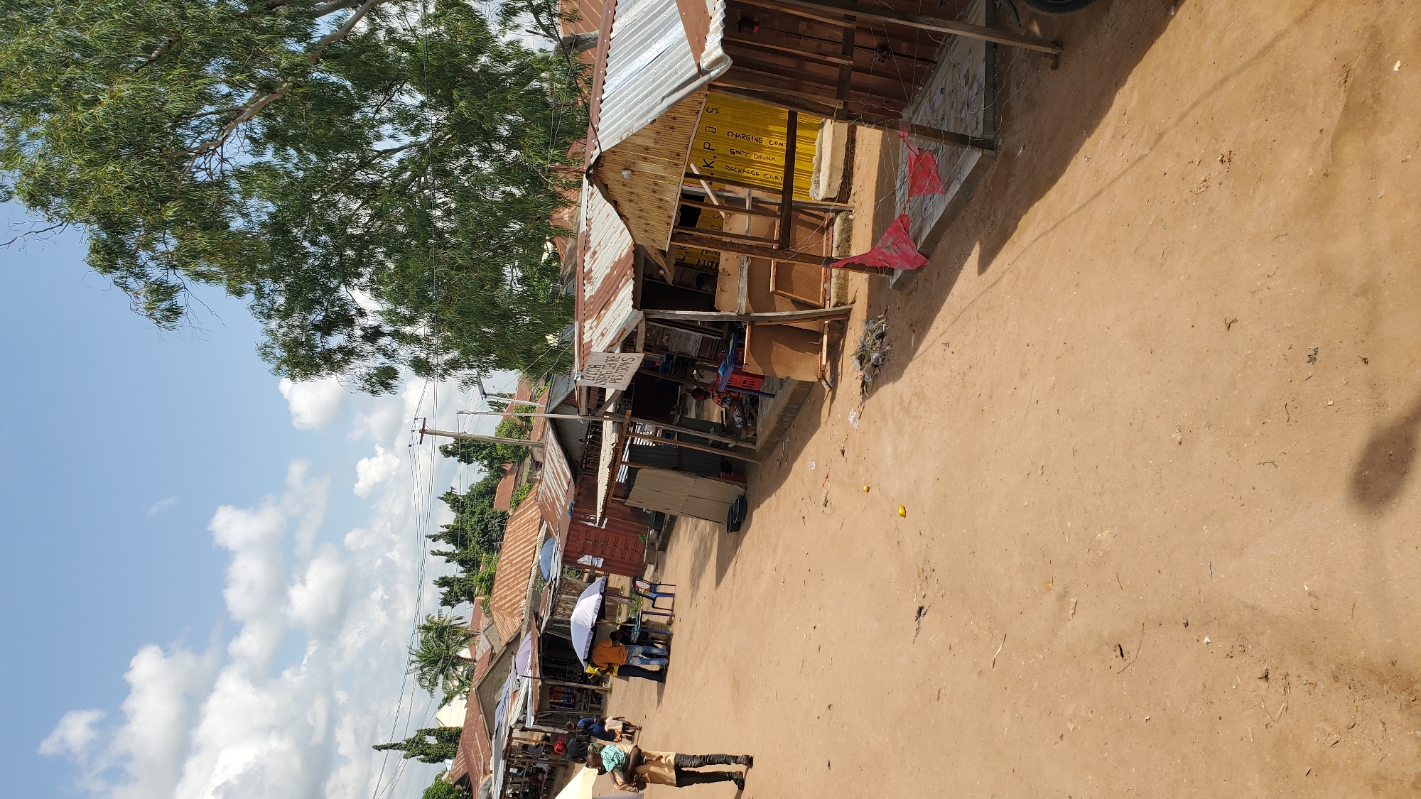 The image shows a street market in Jalingo, Nigeria. The market is lined with small, open-air stalls made of corrugated metal and wood. Some of the stalls have signs, one of which reads "PLUGGED CENTER PICS GRINS." The ground is unpaved and dusty. A few people are visible in the market, including a man carrying a child on his shoulders and several people standing around one of the stalls. Trees and buildings are visible in the background. The sky is blue with scattered white clouds.