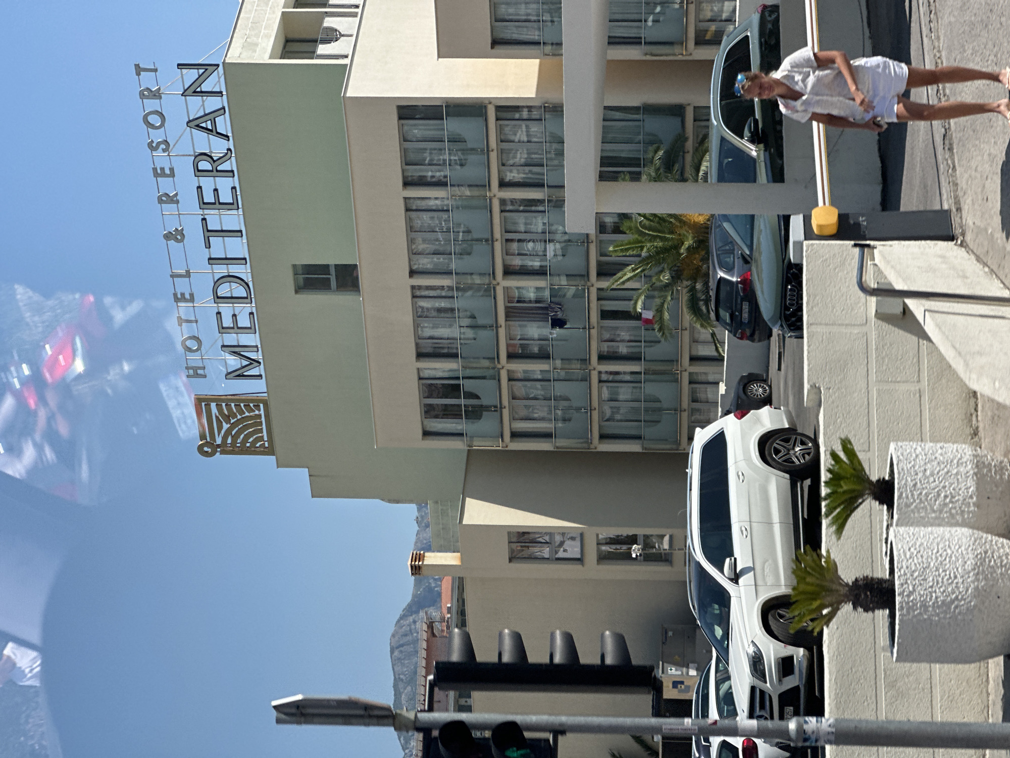 In Boreti, Montenegro, the facade of Hotel & Resort Mediteran is visible on a sunny day. The hotel's signage, featuring the words "HOTEL & RESORT MEDITERAN," is prominently displayed. The building is a multi-story structure with numerous balconies and windows. In the foreground, a white Mercedes-Benz SUV is parked. A woman in white shorts and a white shirt is walking past a barrier. Traffic lights and a traffic signal pole are also present on the left side of the image. The scene suggests a typical urban or resort environment with vehicles, a hotel, and pedestrian activity.