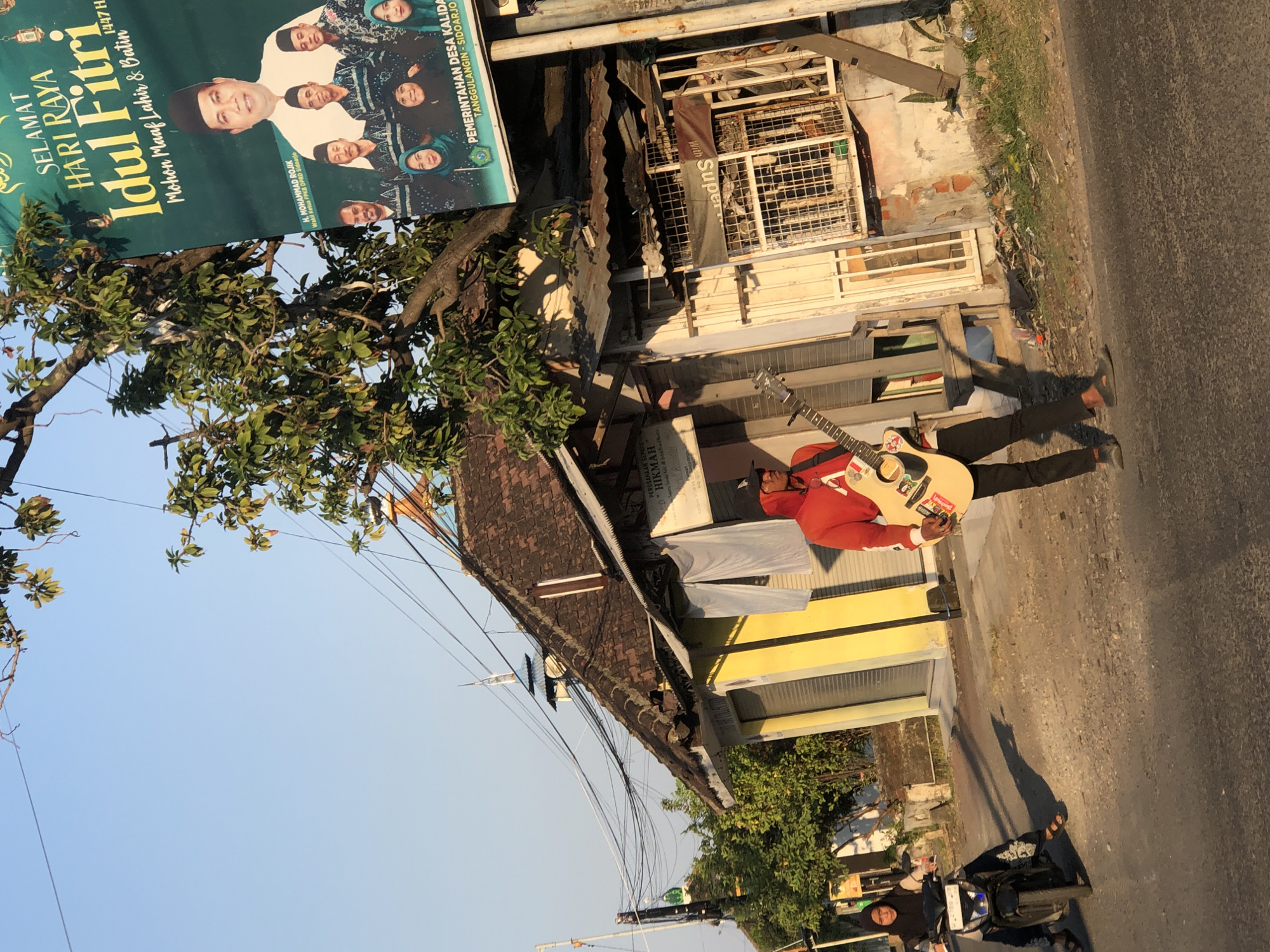 A man in a red hoodie and black pants is playing a guitar on a street in Kalidawir, Indonesia. He is holding a light-colored acoustic guitar and appears to be singing or performing. To the left of the man, a woman wearing a hijab is riding a motorcycle. In the background, there are several small shops with traditional Indonesian architecture, including tiled roofs and wooden structures. A large banner displaying a greeting for Eid al-Fitr, featuring portraits of local politicians and the text "SELAMAT HARI RAYA IDUL FITRI," is visible on the right side of the image. The sky is clear and blue, suggesting it is daytime, likely late afternoon given the long shadows. Power lines are strung across the scene, typical of many Indonesian towns. The environment appears somewhat cluttered, with debris visible along the side of the road.