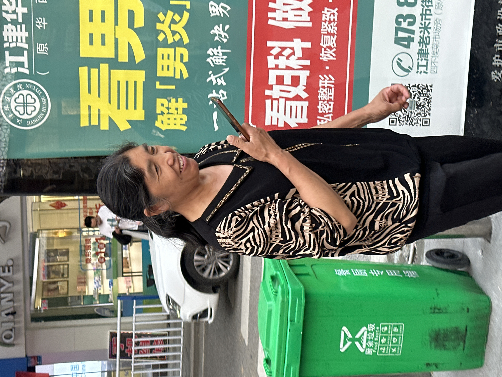 An outdoor shot shows an elderly woman, possibly of Asian descent, looking up and smiling as she holds a smartphone. She is standing next to a green waste bin. In the background is a white car and a store. A large advertisement on the side of a building is in Chinese script. The ad reads "Jiangjin Hua". The location is Jiangjin, China.