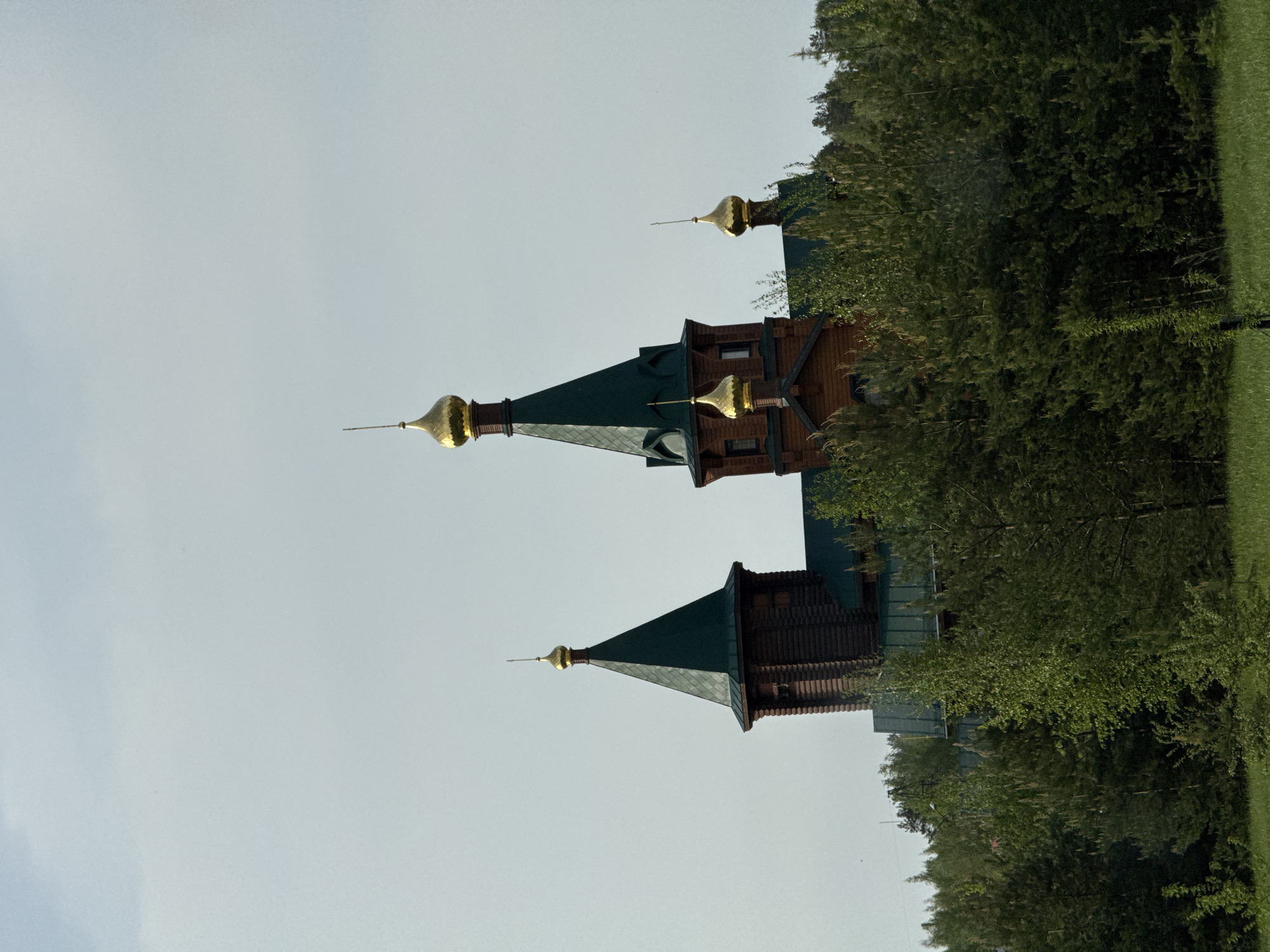 The image shows a wooden church with golden domes in Kstovo, Russia. The building has a dark brown facade and green roofs. The church is partially obscured by trees. The sky is overcast.