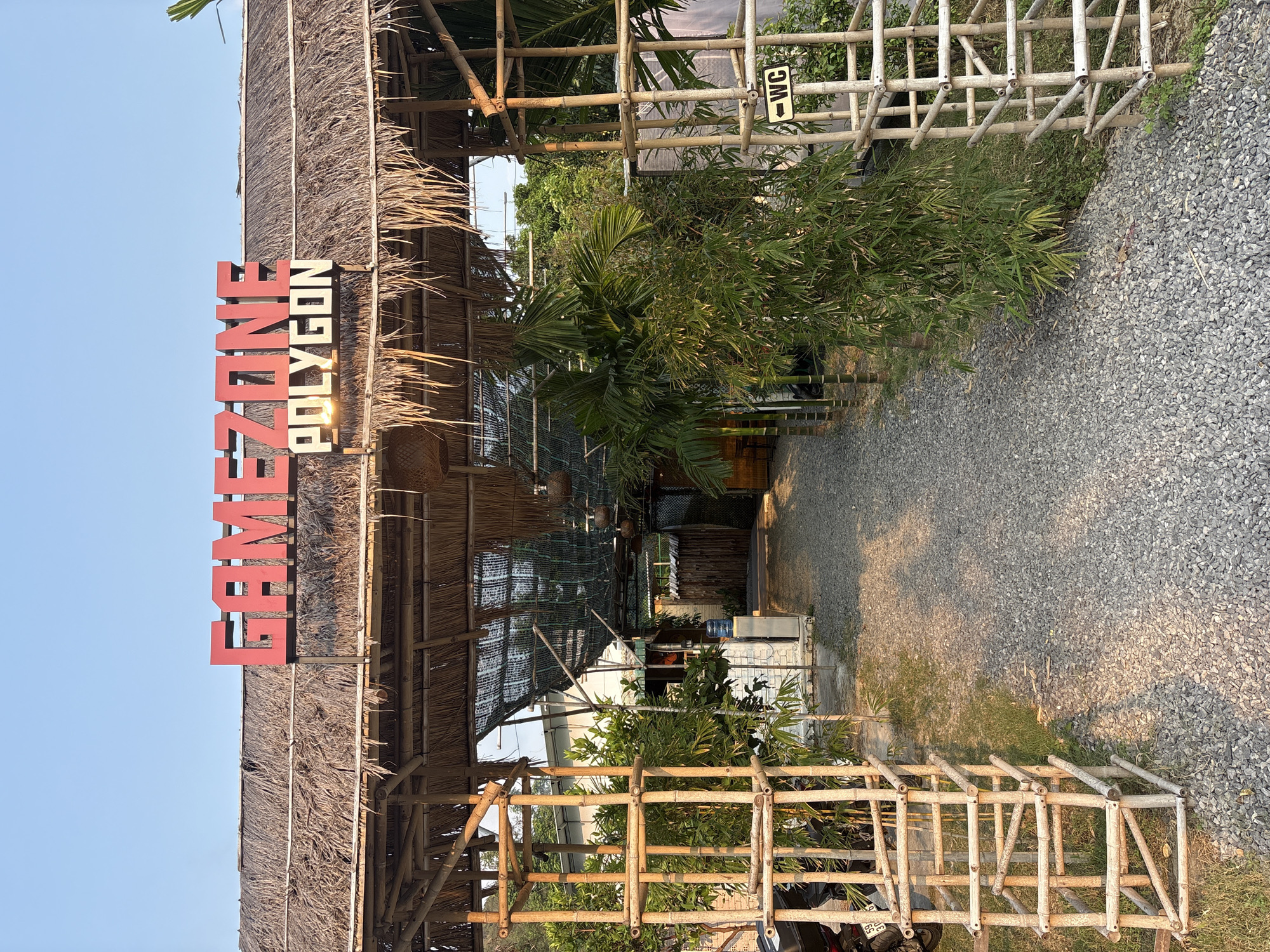 The image depicts the entrance of "GAME ZONE POLYGON" in Bắc Nha Trang, Vietnam. The entrance is marked by a thatched-roof structure with the sign prominently displayed in red. A bamboo fence lines the gravel path leading into the establishment. A WC sign is visible near the entrance. The setting appears to be outdoors, with various plants and trees surrounding the structure. A motorcycle is parked near the fence. The sky is clear and light blue.