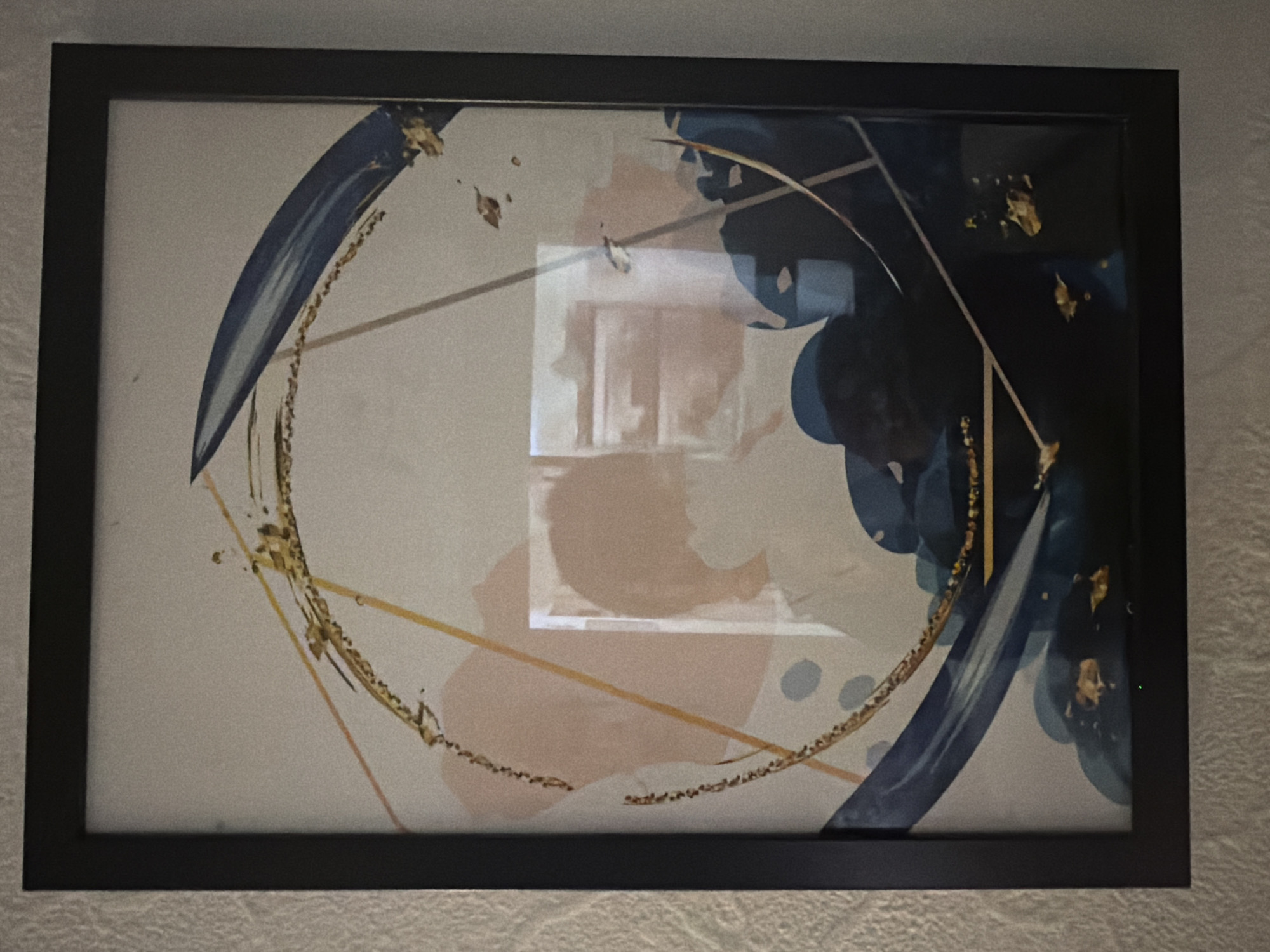 The image shows a framed abstract art print with a dark frame. The artwork features a combination of light and dark blue brushstrokes, golden leaf accents, and geometric lines on a neutral background with blush pink undertones. A circular element is partially depicted with a textured golden border. A square reflection is visible in the center of the print, showing a portion of an interior room with vertical architectural elements. The print is mounted on a textured, light-colored wall. The photograph does not contain any information pertaining to the city of London.