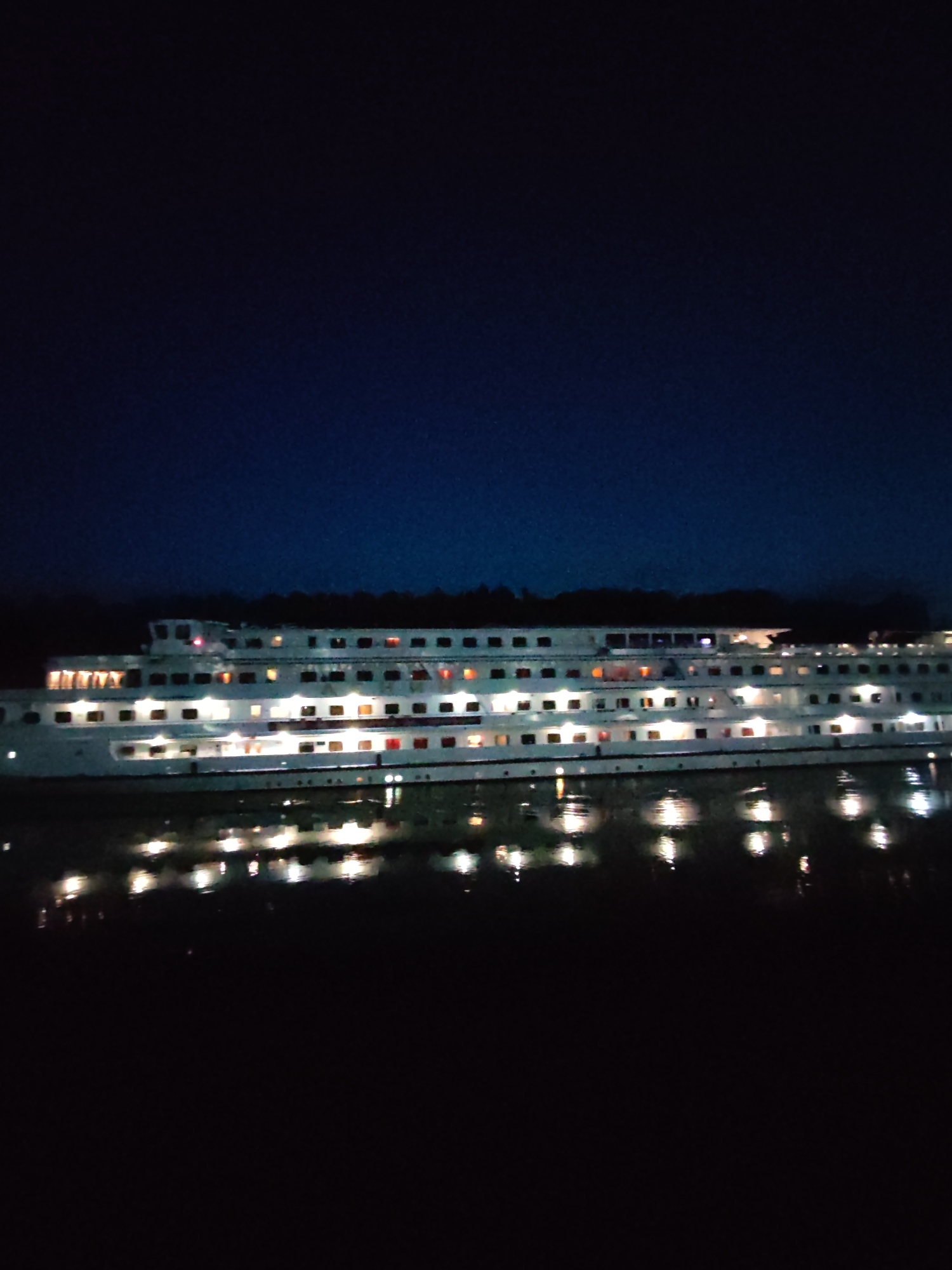 An illuminated white cruise ship is docked at night in the city of Rybinsk, Russia. The ship has multiple decks with rows of lit windows reflecting on the dark water it is docked in. The sky above is a deep blue, fading to black at the top of the image. The reflections of the ship's lights create a shimmering effect on the water's surface.