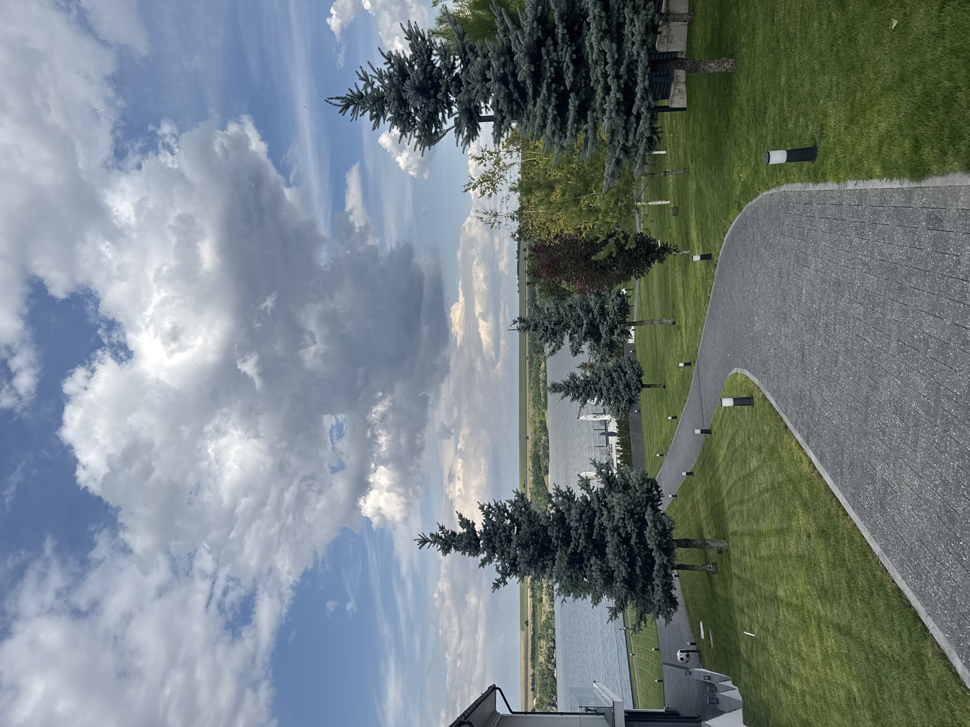 The image depicts a scenic view from a property in Oxentea, Moldova. In the foreground, a winding paved pathway made of dark grey interlocking stones leads through a well-manicured green lawn. The lawn is bordered by a concrete edge and is dotted with several modern, black and white bollard lights. Several mature fir trees with dark green foliage are strategically placed along the pathway and on the lawn.
To the left, a section of a modern white building with dark grey accents is visible, featuring a balcony with glass railings. Beyond the lawn and trees, a wide body of water, likely a river or lake, stretches across the mid-ground. The water is calm and reflects the sky. On the opposite bank of the water, a gently sloping landscape of green fields and trees is visible, leading to a horizon under a dramatic sky.
The sky is a prominent feature, filled with a mix of bright blue patches and large, billowing grey and white clouds, suggesting a dynamic weather system. A few scattered trees, including one with red-tinged leaves, are visible in the middle distance, adding variety to the landscape. The overall impression is one of tranquility and natural beauty, enhanced by the presence of the modern architecture.