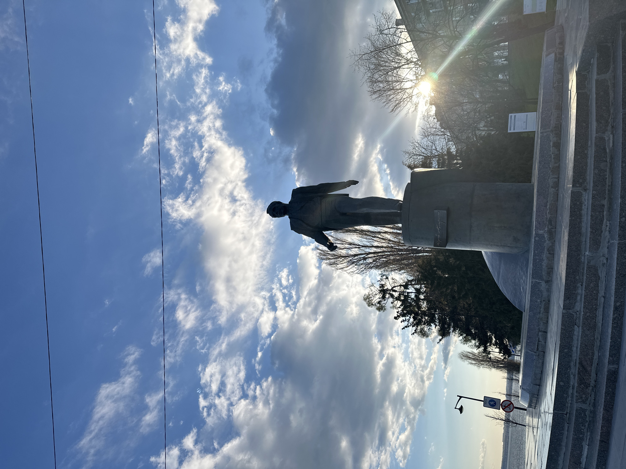 The image depicts an outdoor scene centered on a statue, dramatically backlit by the sun. The main subject is a bronze-colored statue of a man, silhouetted against the sky and standing on a cylindrical stone pedestal. The figure appears to be wearing a suit or uniform, with one arm slightly outstretched.
The sky is a prominent feature, filled with a mix of bright blue areas and large, billowy white and grey clouds. The sun, partially visible in the upper right quadrant behind the statue, creates a strong lens flare, indicating either early morning or late afternoon. Bare tree branches, suggesting a dormant season, are scattered across the upper right, with denser evergreen trees lower down.
To the right of the statue, a flight of granite steps leads upwards. In the background, a building is partially visible, along with a couple of white informational signs whose text is illegible. Further in the distance, a lamppost stands next to a road or pathway. A visible street sign attached to the lamppost clearly shows a "no pedestrians" symbol (a white figure crossed out by a red circle and diagonal line). Thin power lines run across the sky in the upper left. The scene is static, featuring a monument in an urban or park setting. There is no specific activity or event taking place. Visible text on the statue's base and other signs in the background is unreadable due to distance and lighting conditions.