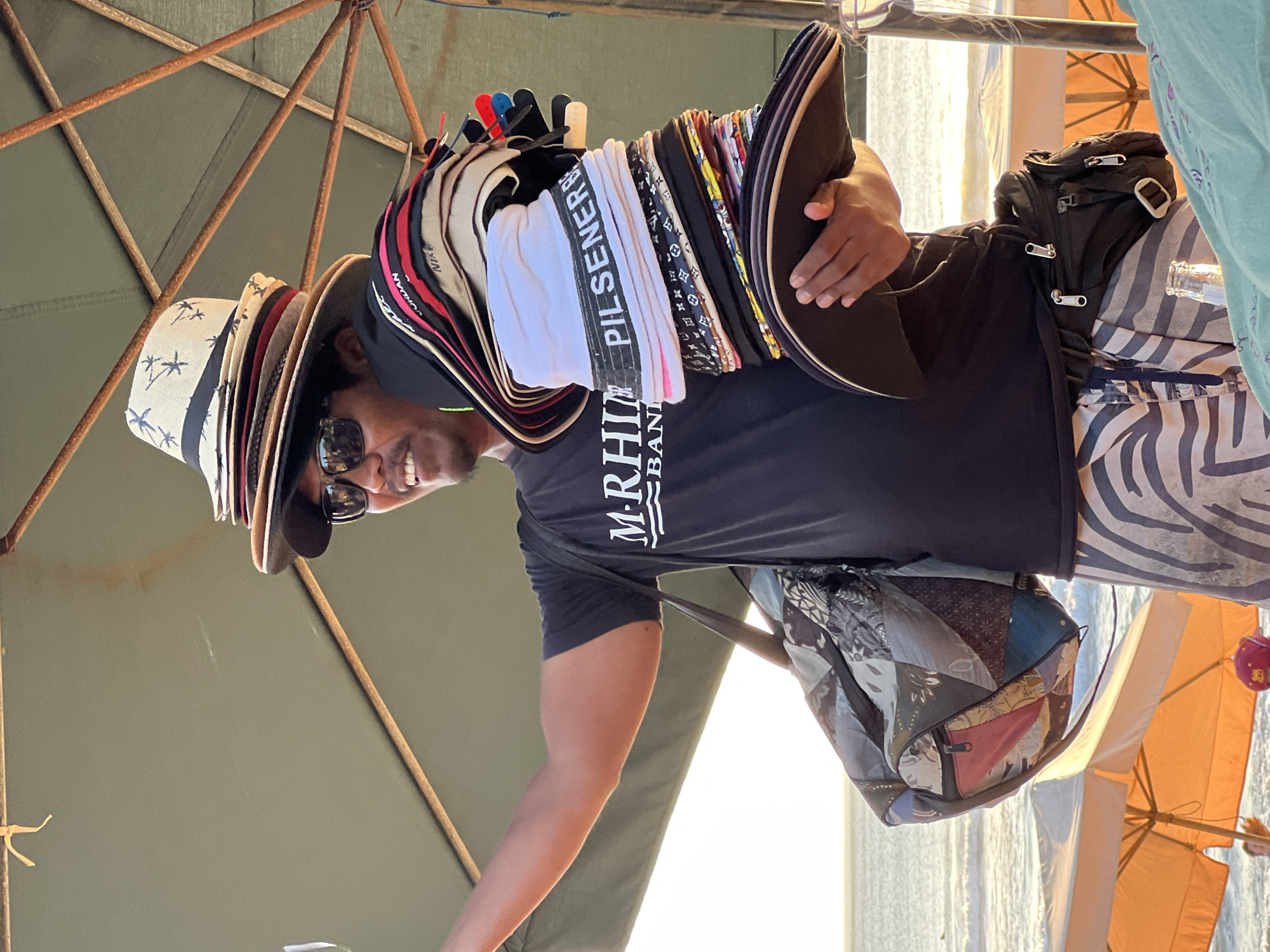 A man is standing under an umbrella at what appears to be a beach or waterfront location, likely Kuta, Indonesia, given the context of the question. He is wearing sunglasses, a black t-shirt with "M-RHINO BAND" printed on it, and patterned shorts. The man is holding a large stack of hats and caps in his right arm, with a duffel bag slung over his left shoulder. The hats are of various styles and colors, with some featuring brand names like "NIKE" and "JORDAN." In the background, there are beach umbrellas and the ocean, suggesting a sunny, outdoor environment. A fanny pack is worn around his waist. The lighting and atmosphere suggest it is late afternoon or early evening.