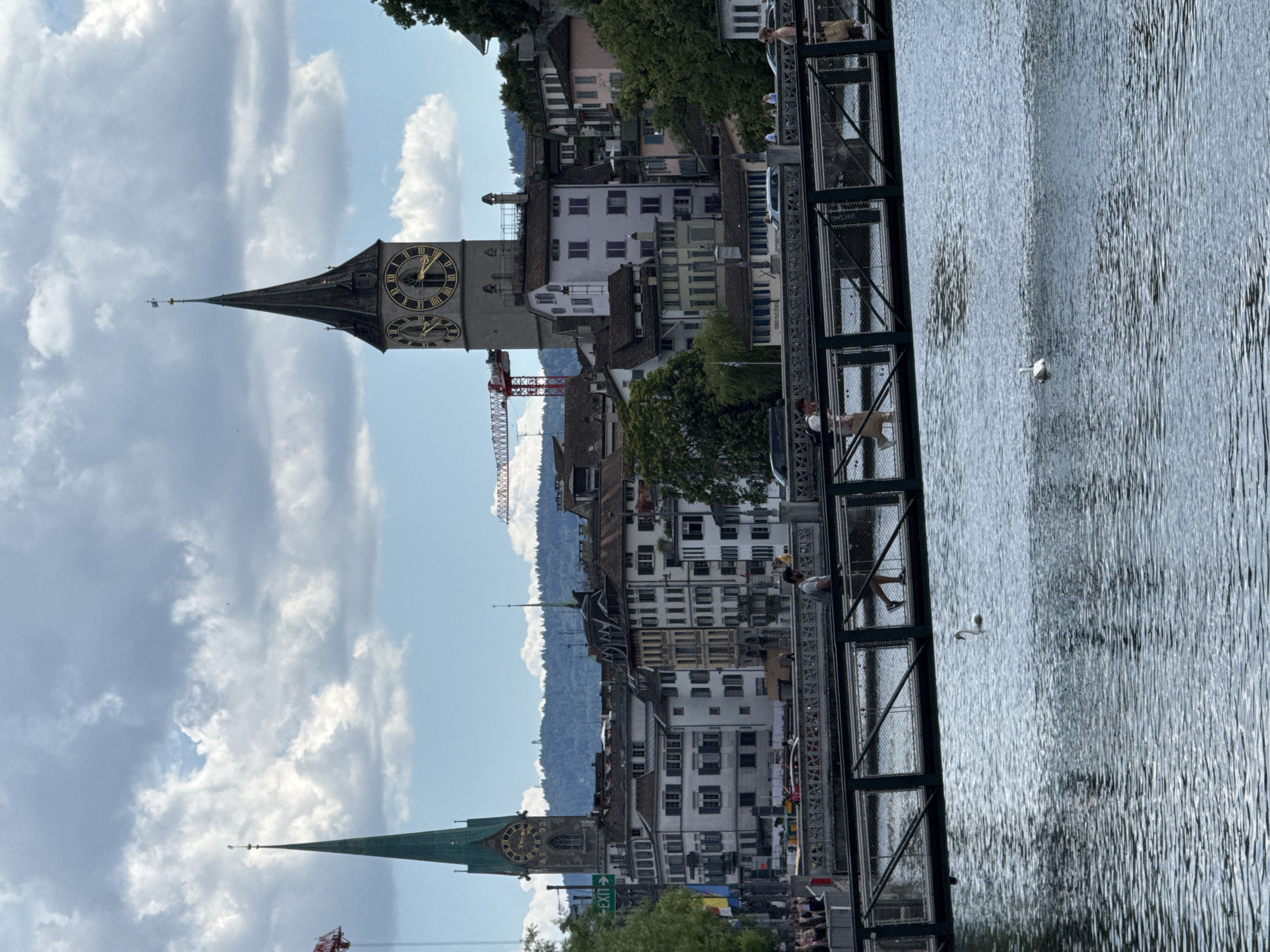 The image depicts a city scene in Zurich, Switzerland, likely along the Limmat River. In the background, two prominent church spires dominate the skyline: one is a tall, green, Gothic-style spire on the left, and the other is a larger, grey clock tower with Roman numerals on its face on the right. Numerous buildings with traditional European architecture, featuring tiled roofs and numerous windows, are clustered between and around the spires. A red construction crane is visible in the middle distance, suggesting ongoing development.
In the foreground, a modern pedestrian bridge spans the water. Several individuals are walking across the bridge, some of whom appear to be tourists. A swan is swimming in the calm, blue water below the bridge, its reflection visible on the rippling surface. The sky is partly cloudy, with a mix of white and grey clouds against a bright blue backdrop. The overall impression is of a vibrant urban environment with a blend of historical and modern elements.