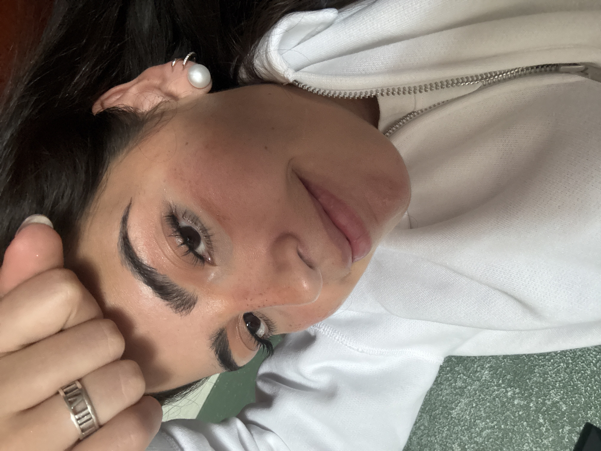 A close-up, slightly rotated image of a woman wearing a white hooded sweatshirt and silver jewelry, possibly lying down. Her face is prominent, with dark hair, defined eyebrows, and visible eyelashes. She wears a ring on her finger and earrings in her ear. The background appears to be a textured surface. There is no evidence to specify the city of San Antonio, United States of America.