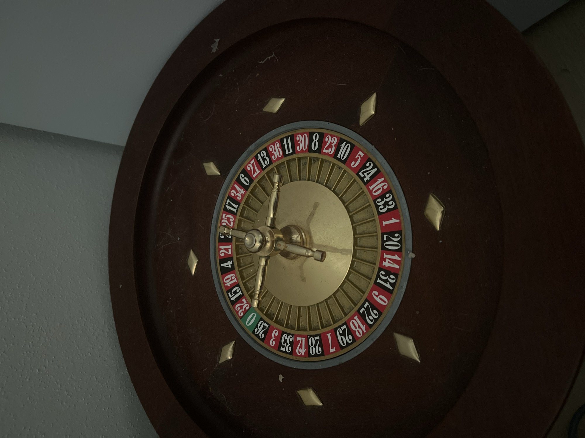 The image shows a close-up, overhead view of a roulette wheel. The wheel is made of dark wood with gold diamond-shaped inlays around the rim. The numbers on the roulette wheel are visible and appear to be in a standard European or American layout, with red and black pockets and a green zero pocket. The center of the wheel has a gold-colored mechanism with spokes. Dust particles are visible on the surface of the wheel and the surrounding wood. The background consists of a textured, light-colored wall and a darker, angled surface. The lighting is dim, casting shadows and highlighting the details of the roulette wheel. There are no people or animals present in the image, and no specific location in Acquaviva, Italy, can be determined from the visual information provided.