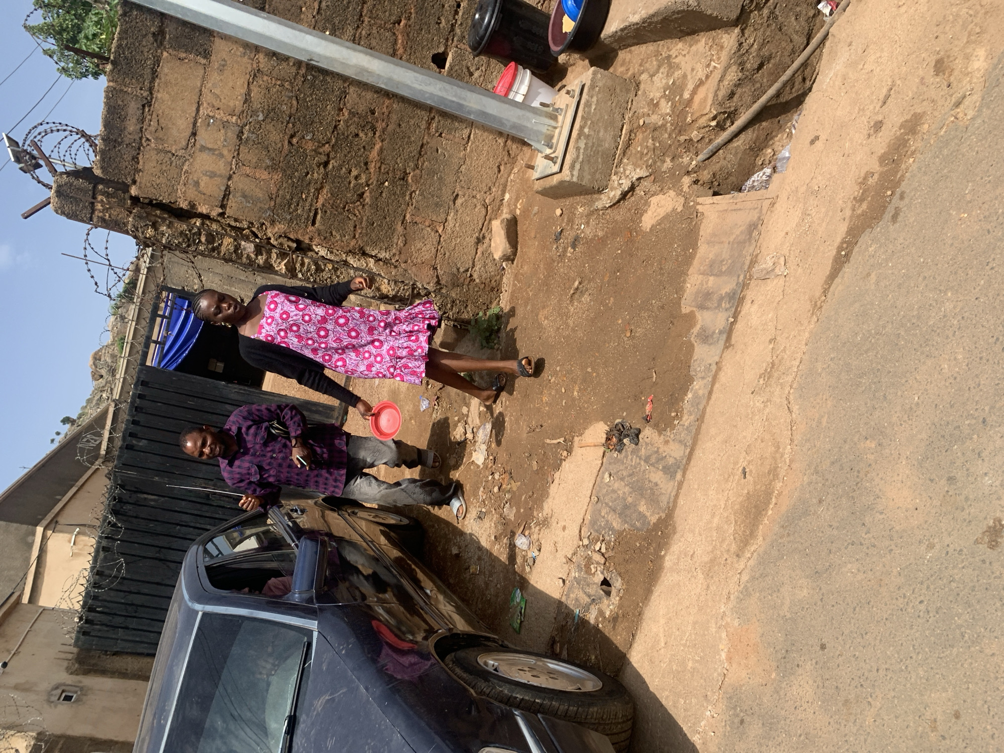 Two individuals stand near a dark-colored sedan on a street. A man in a purple checkered shirt is next to the car. A woman in a pink dress stands slightly behind him. A red plastic plate is in her hand. The car is parked near a metal gate topped with barbed wire. The street is paved, with debris and dirt along the edge. A brick wall borders the street. A utility pole is visible next to the wall. This scene is in a city.