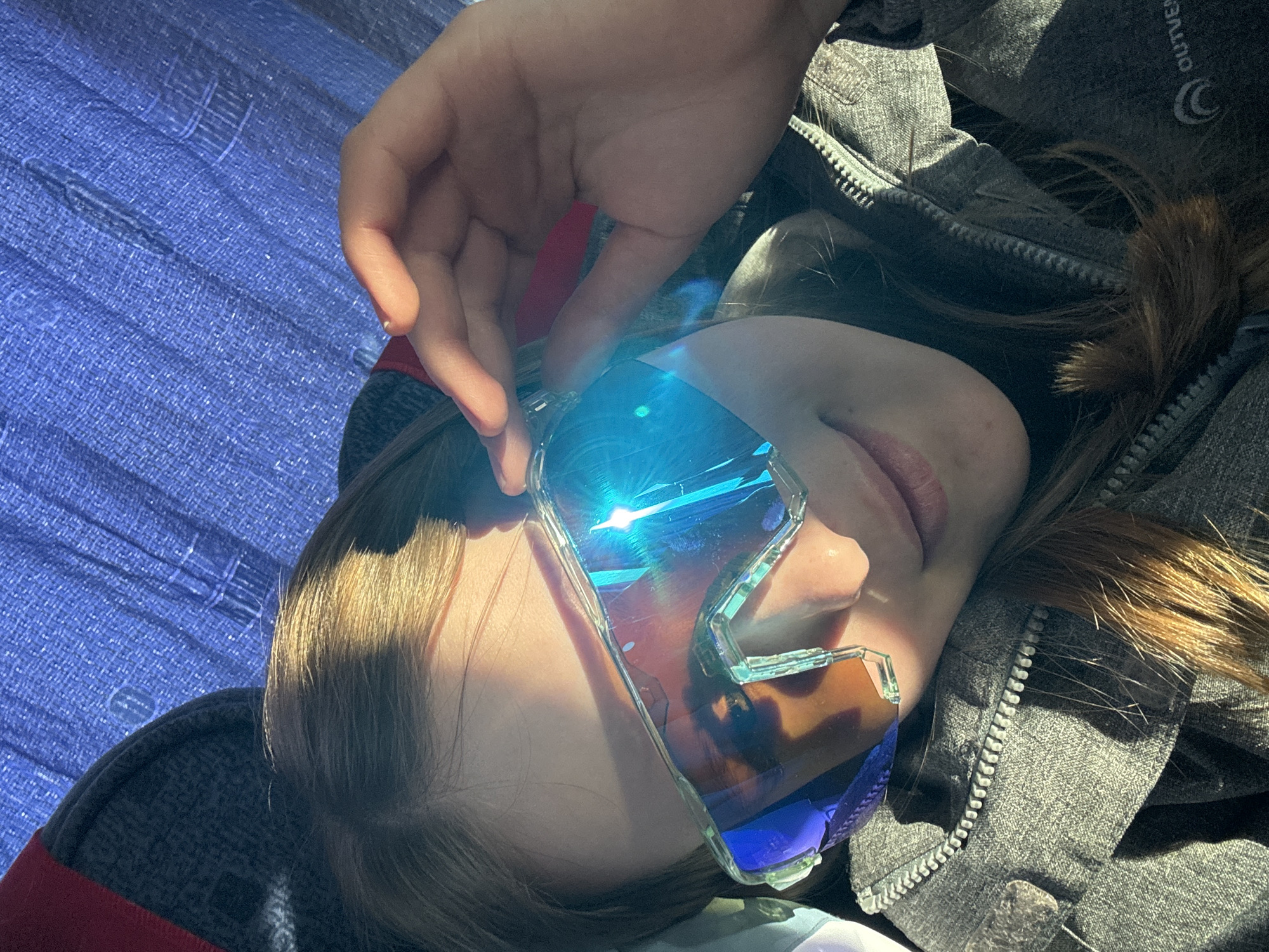 A person wearing sunglasses is depicted in a close-up, slightly angled shot. The sunglasses have blue lenses and a clear frame. The person's hand is visible, holding the glasses in place. The background includes a gray jacket and blue textured material. The image was taken outdoors in daylight. It is impossible to confirm the location is the city of Berezniki, Russia, without more information or context.