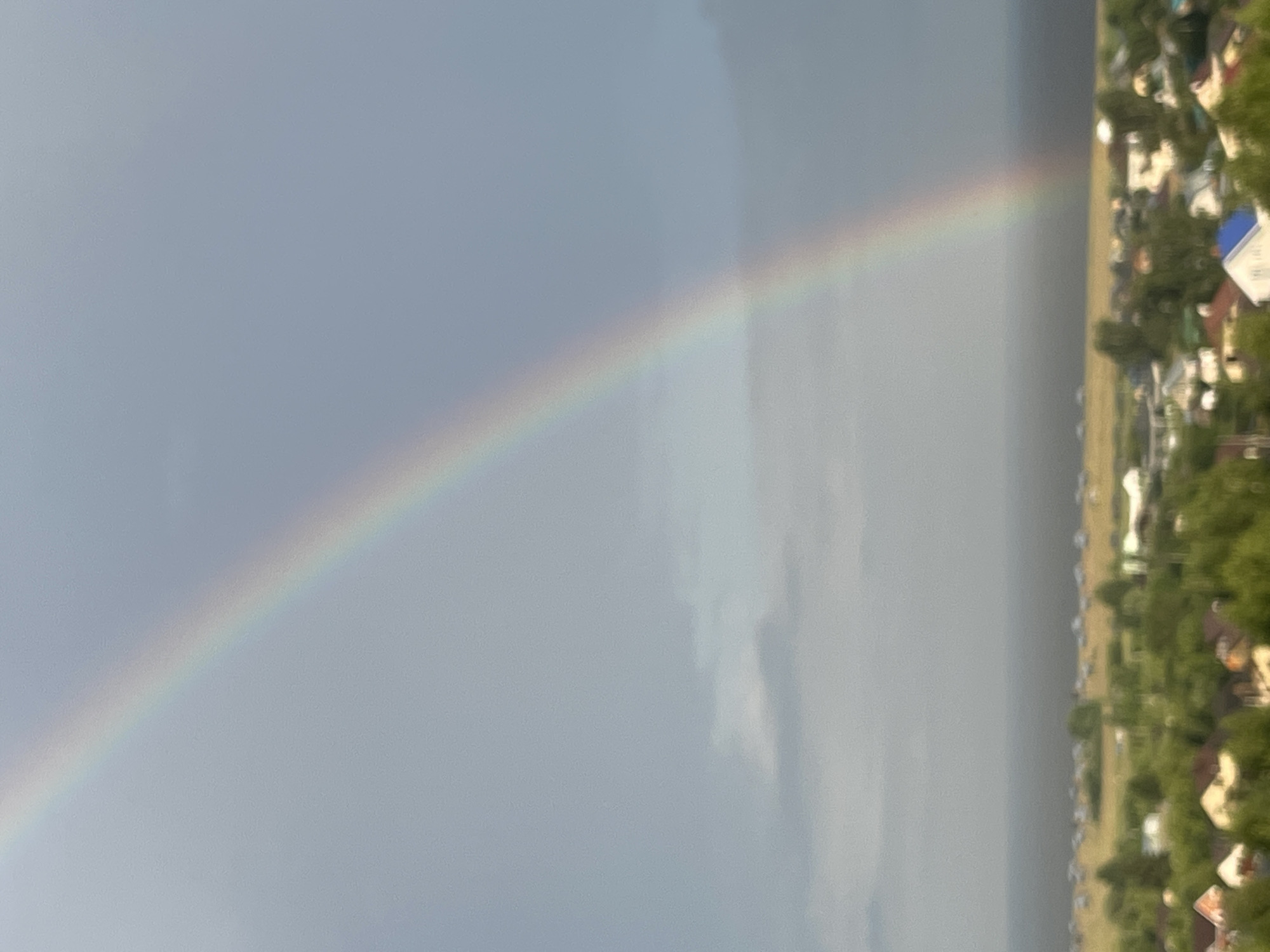 A rainbow arcs across a grey, cloudy sky over a rural landscape. In the foreground, a cluster of houses with red and white roofs are nestled among green trees. A body of water is visible in the middle ground, and beyond that, a flat expanse of land with scattered structures. The image appears to be taken from an elevated position, offering a panoramic view of the area, consistent with a location in Laishevo, Russia.