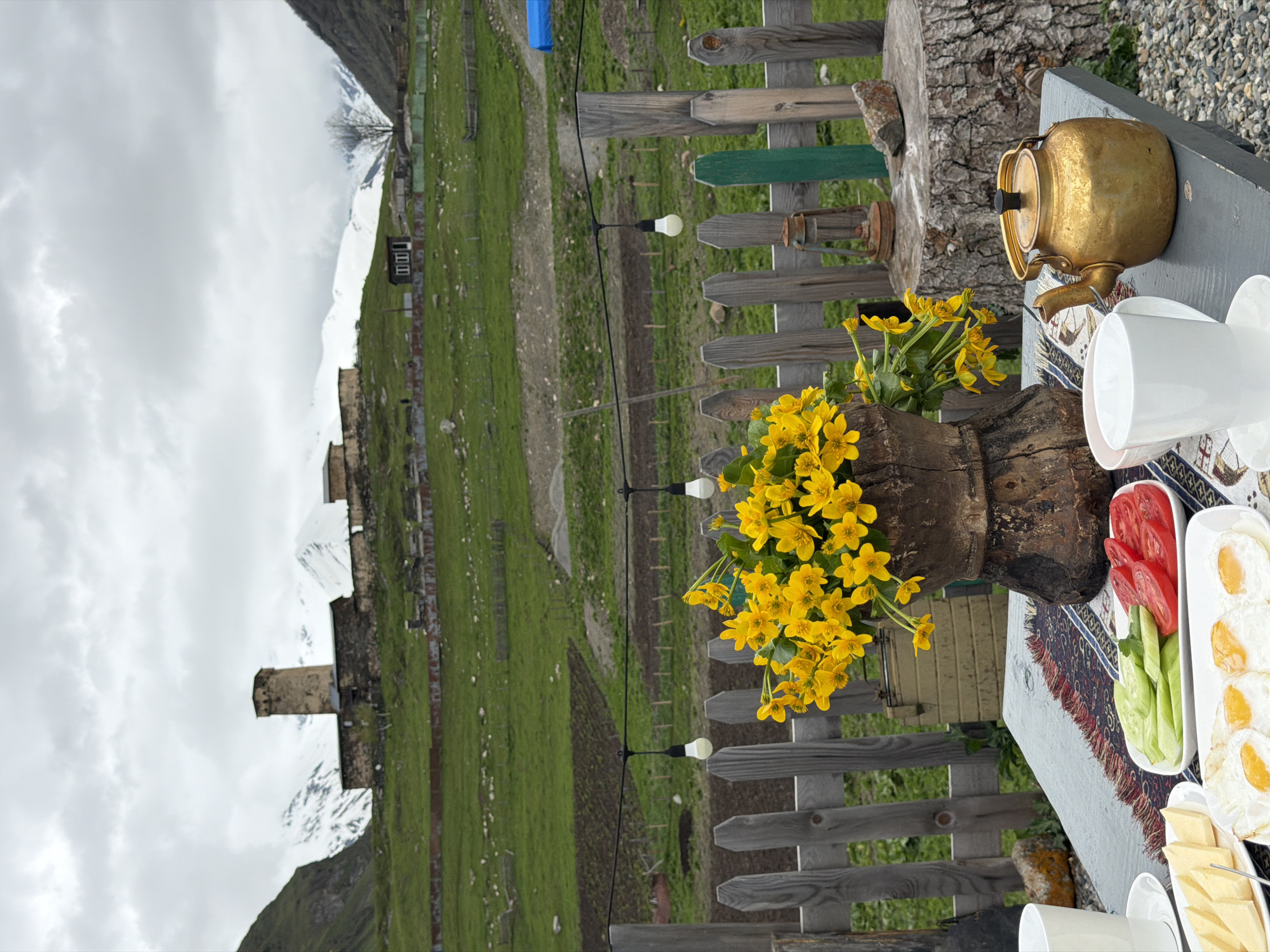 In Ushguli, Georgia, a table is set for breakfast outdoors. In the foreground, a golden teapot, white cups, and plates of food, including fried eggs, cheese, sliced tomatoes, and cucumbers, are arranged on a table adorned with a patterned runner. A rustic wooden vase overflowing with bright yellow flowers sits on the table. Behind the table, a weathered wooden fence encloses the scene, with an old lantern resting on a tree stump. In the background, a lush green hillside slopes upwards, dotted with agricultural fields and traditional Georgian stone towers, some of which appear to be part of a fortified village. Snow-capped mountains loom in the distance under a cloudy sky, completing the picturesque mountain landscape.